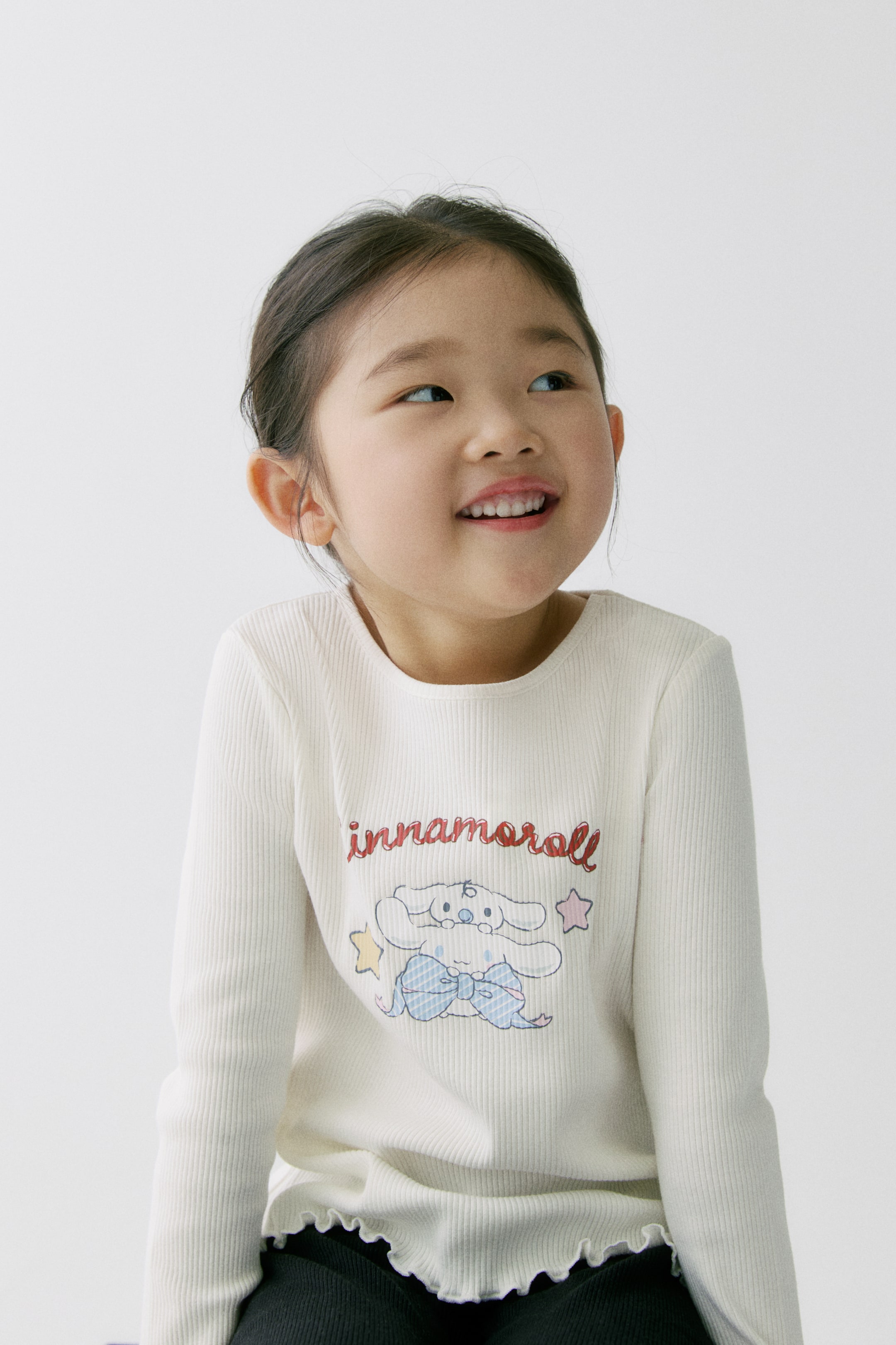 이미지 확대 보기: 크림색 긴소매 골지 탑은 "cinnamoroll" 그래픽이 프린트되어 있으며, 만화 캐릭터와 별을 특징으로 하고 라운드 넥과 물결 모양의 밑단으로 디자인되었습니다.