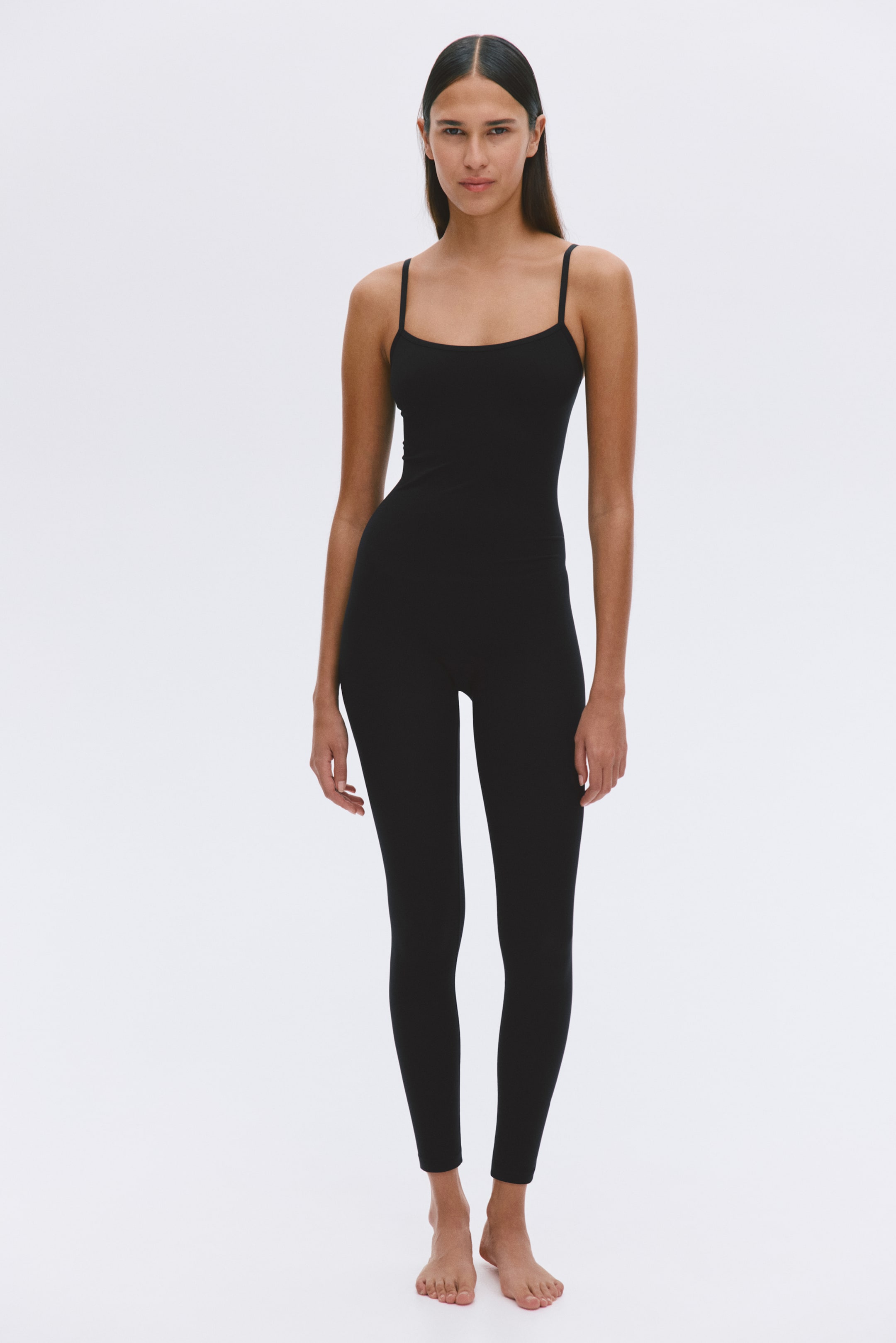 Grotere afbeelding bekijken: De model draagt een zwarte unitard van stretchjersey. Het nauwsluitende kledingstuk heeft een rechte halslijn, smalle schouderbandjes en pijpen tot aan de enkels.