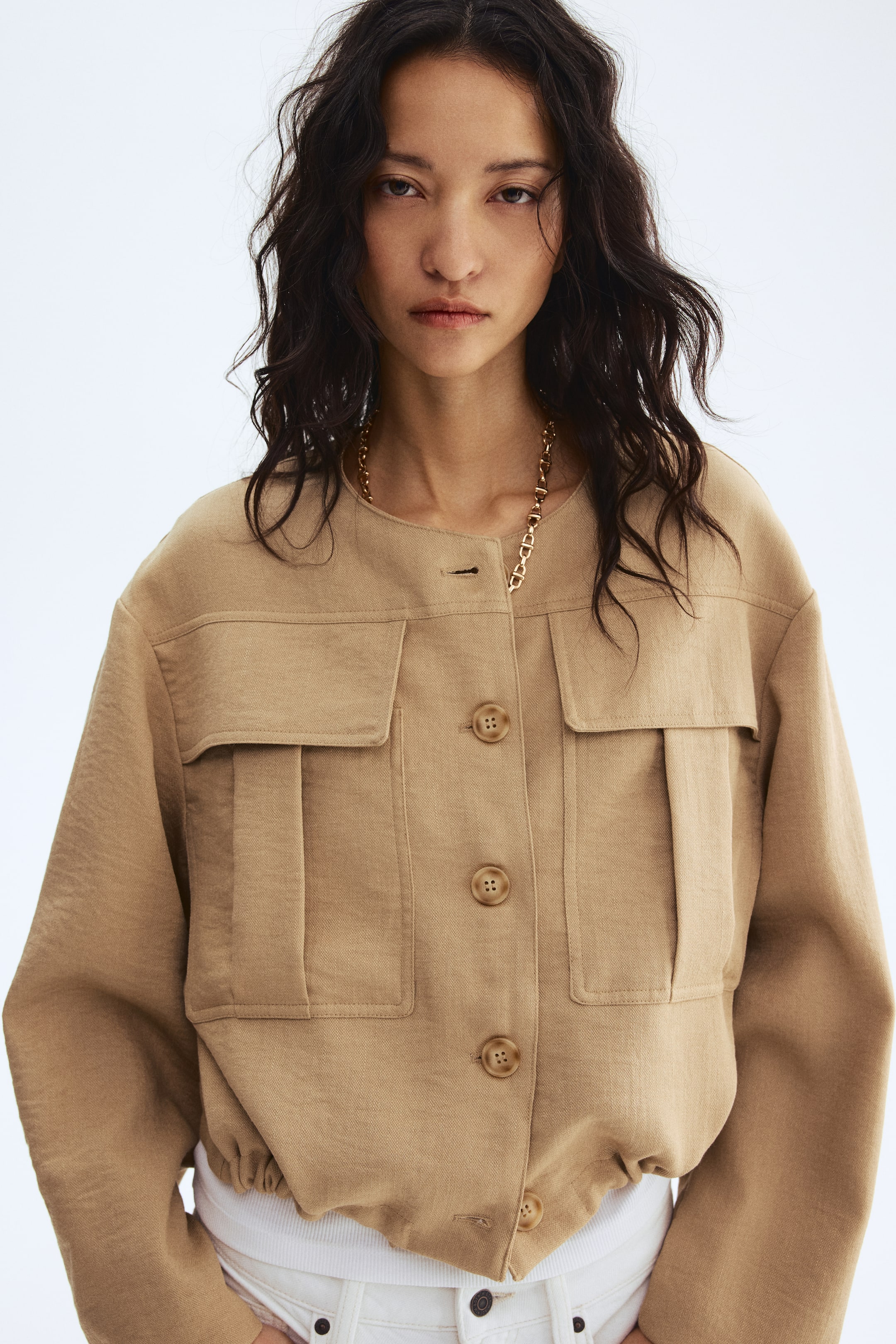 View larger image: A light brown cropped jacket featuring a round collarless neckline and a button-front closure with visible buttons. Designed with two large pleated flap pockets on the chest, an elasticised hem, and a white garment visible underneath.