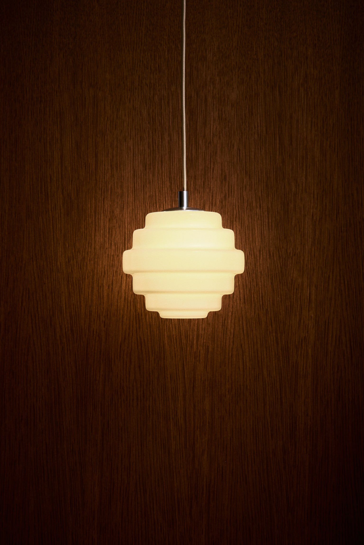 A pendant lamp viewed from the front is hanging from a thin, fabric-covered cord. It features a tiered geometric glass lampshade, emitting a warm, soft light from within. - 1