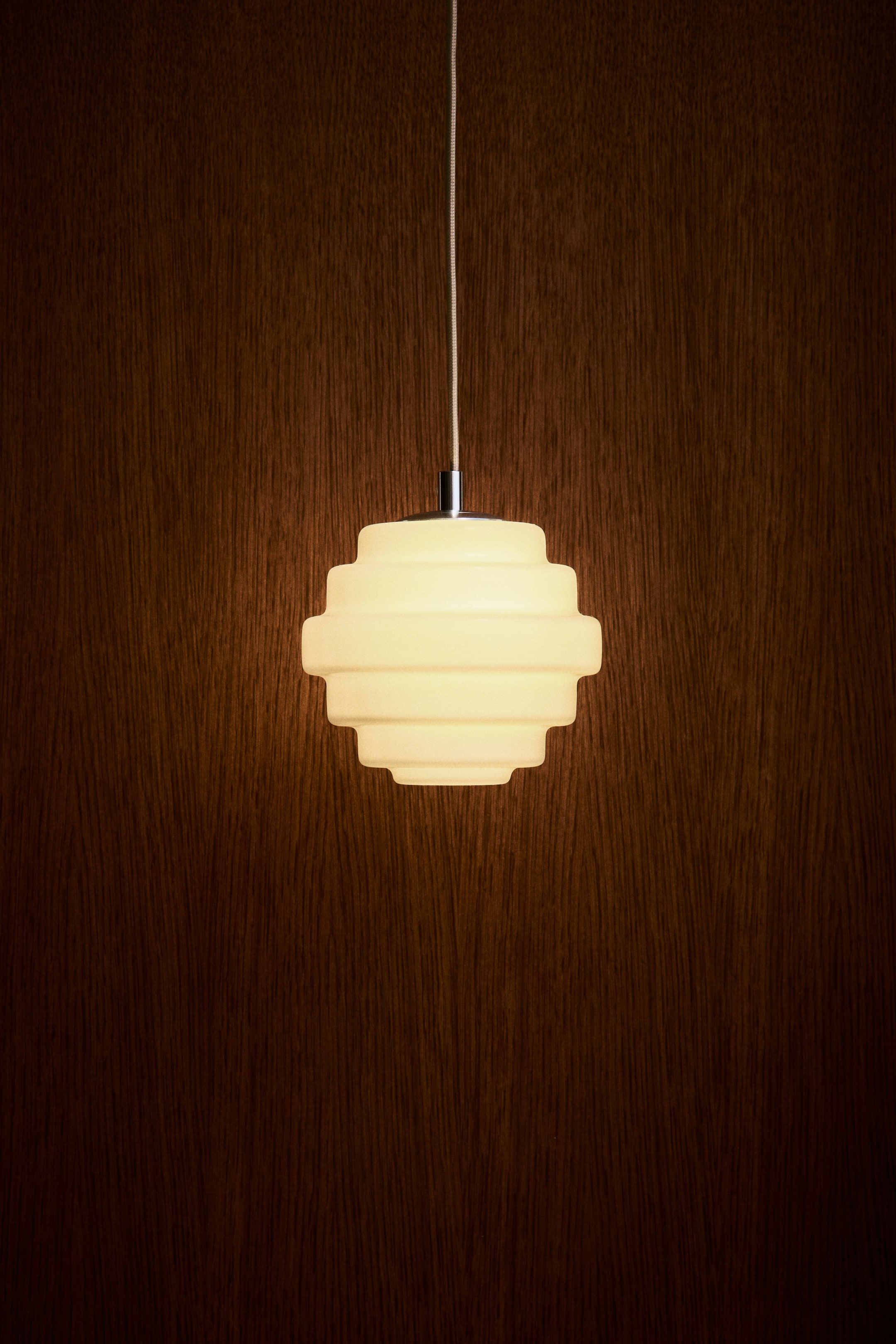 Agrandir l'image: Une lampe suspendue vue de face est accrochée à un fin cordon gainé de tissu. Elle présente un abat-jour en verre géométrique à étages, diffusant de l’intérieur une lumière chaude et douce.