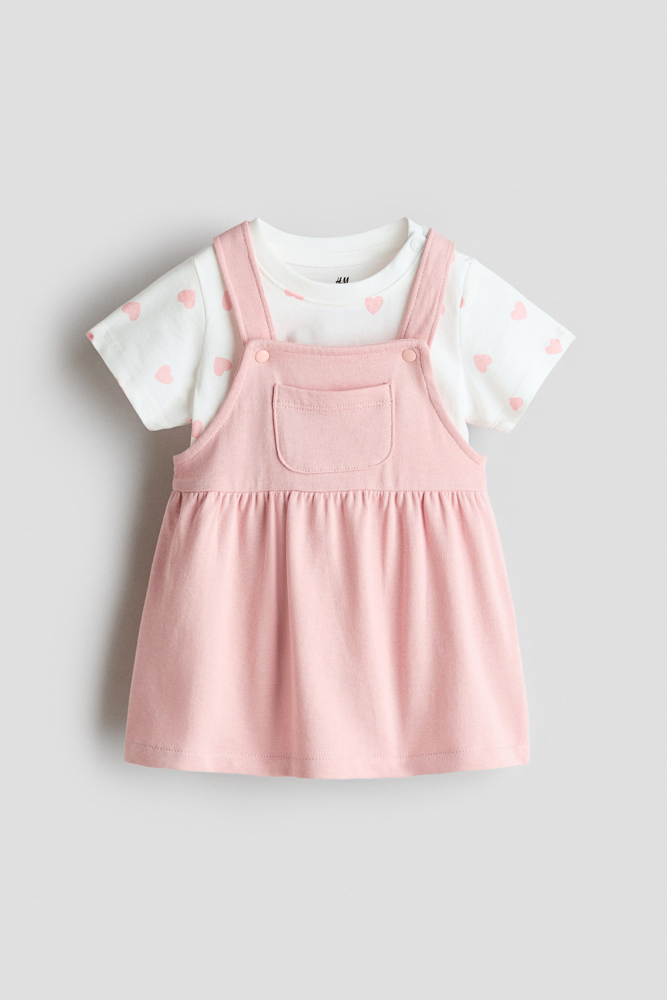 View larger image: A baby's two-piece set is shown facing forward, featuring a white short-sleeved top patterned with small pink hearts layered beneath a pink overall dress. The overall presents a front bib with a patch pocket, and its shoulder straps fasten with buttons above a skirt gathered at the waist.