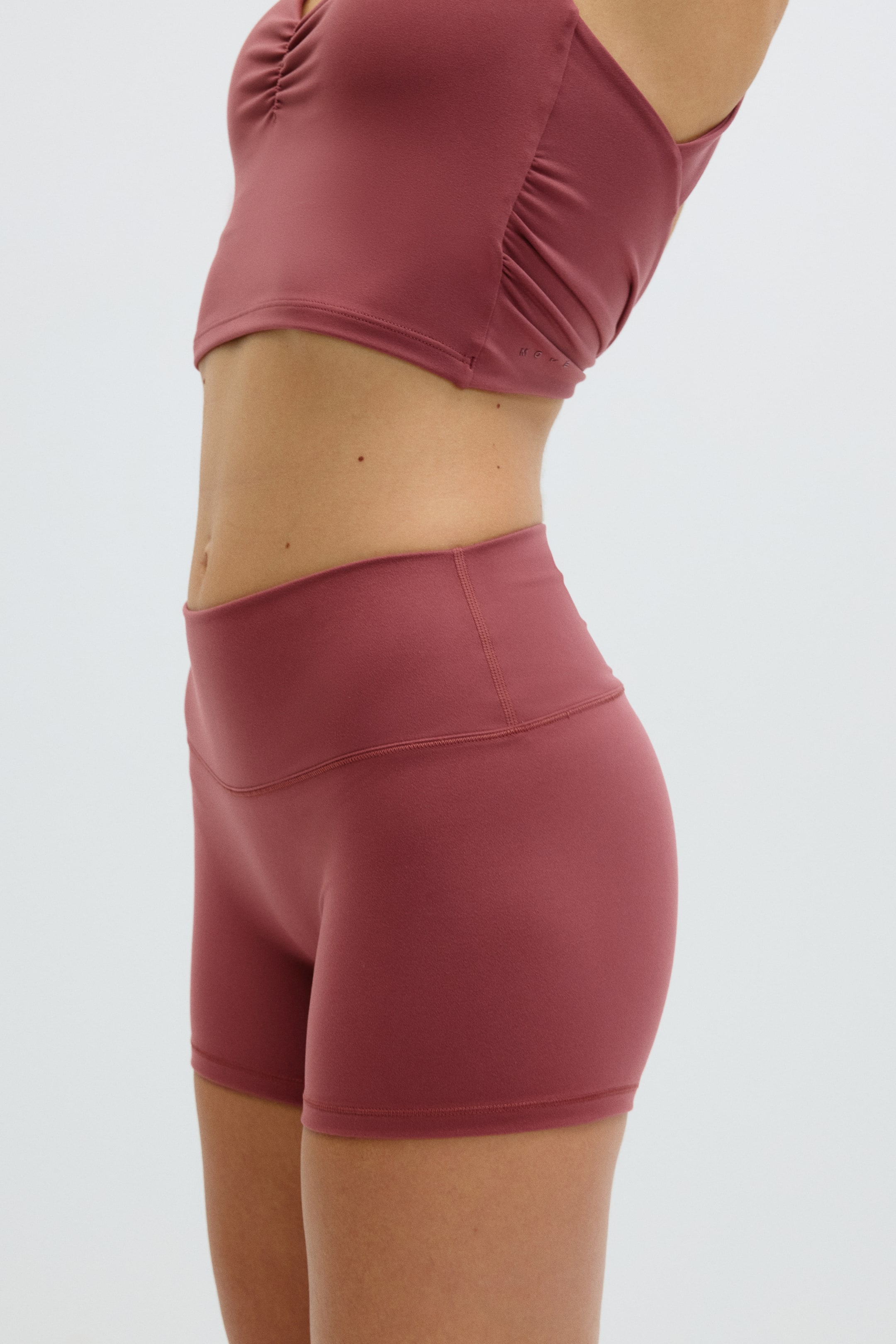 Ver imagen más grande: "Un conjunto de ropa deportiva en color rosa empolvado se muestra desde un ángulo lateral, con un top corto que presenta fruncidos en los laterales y pantalones cortos de tiro alto. Los pantalones cortos tienen un diseño ajustado con una cintura ancha."