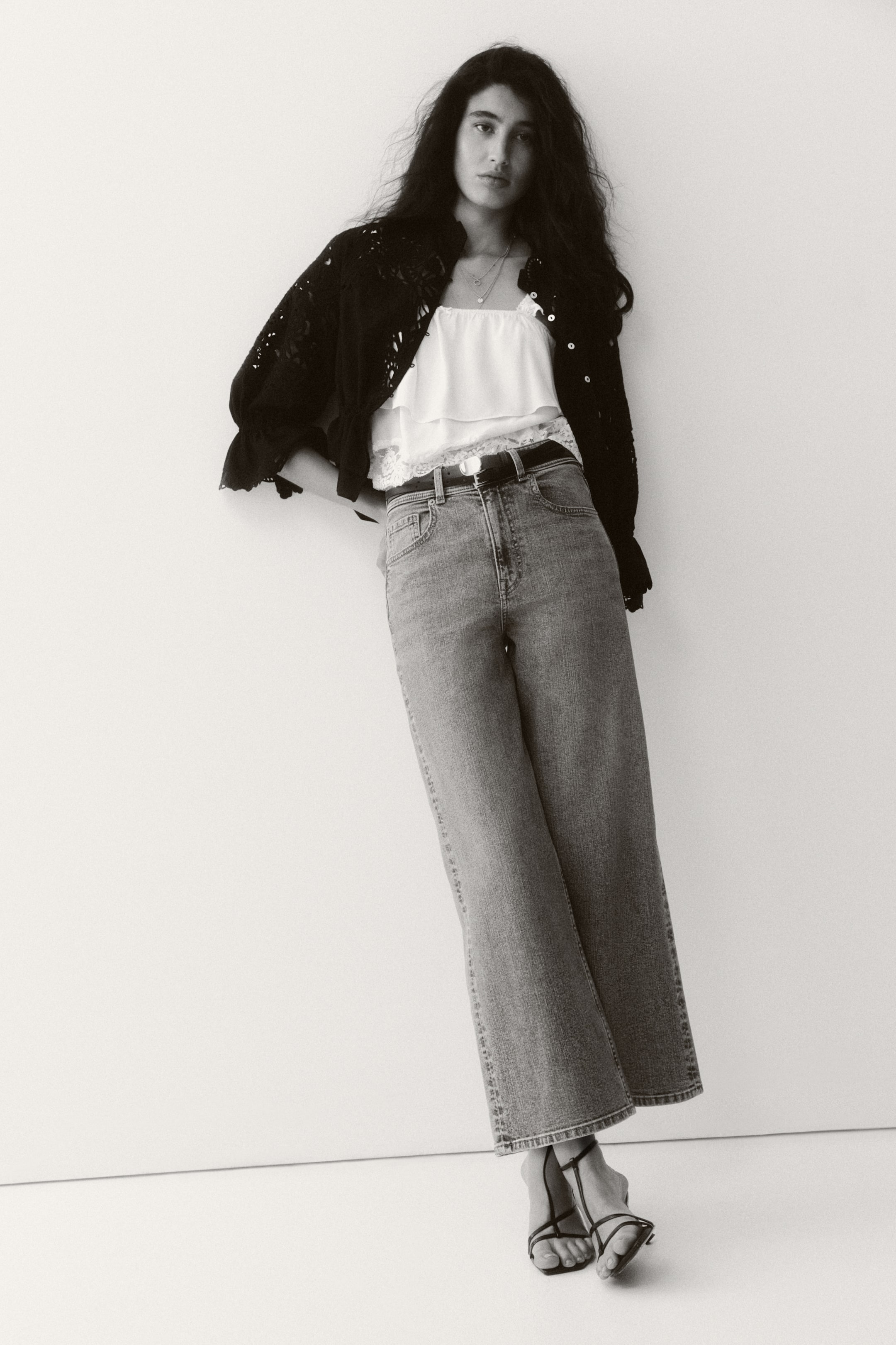 Größeres Bild ansehen: Eine schwarze, offen gestrickte Jacke wird offen über einem weißen Rüschen-Top mit Spitzenbesatz getragen, und zwei silberfarbene Halsketten schmücken den Hals. Hell gewaschene, hoch taillierte Jeans mit weitem Bein werden aus einer frontalen Perspektive präsentiert, durch einen schwarzen Gürtel gebunden, und schwarze Riemchensandalen vervollständigen den Look.