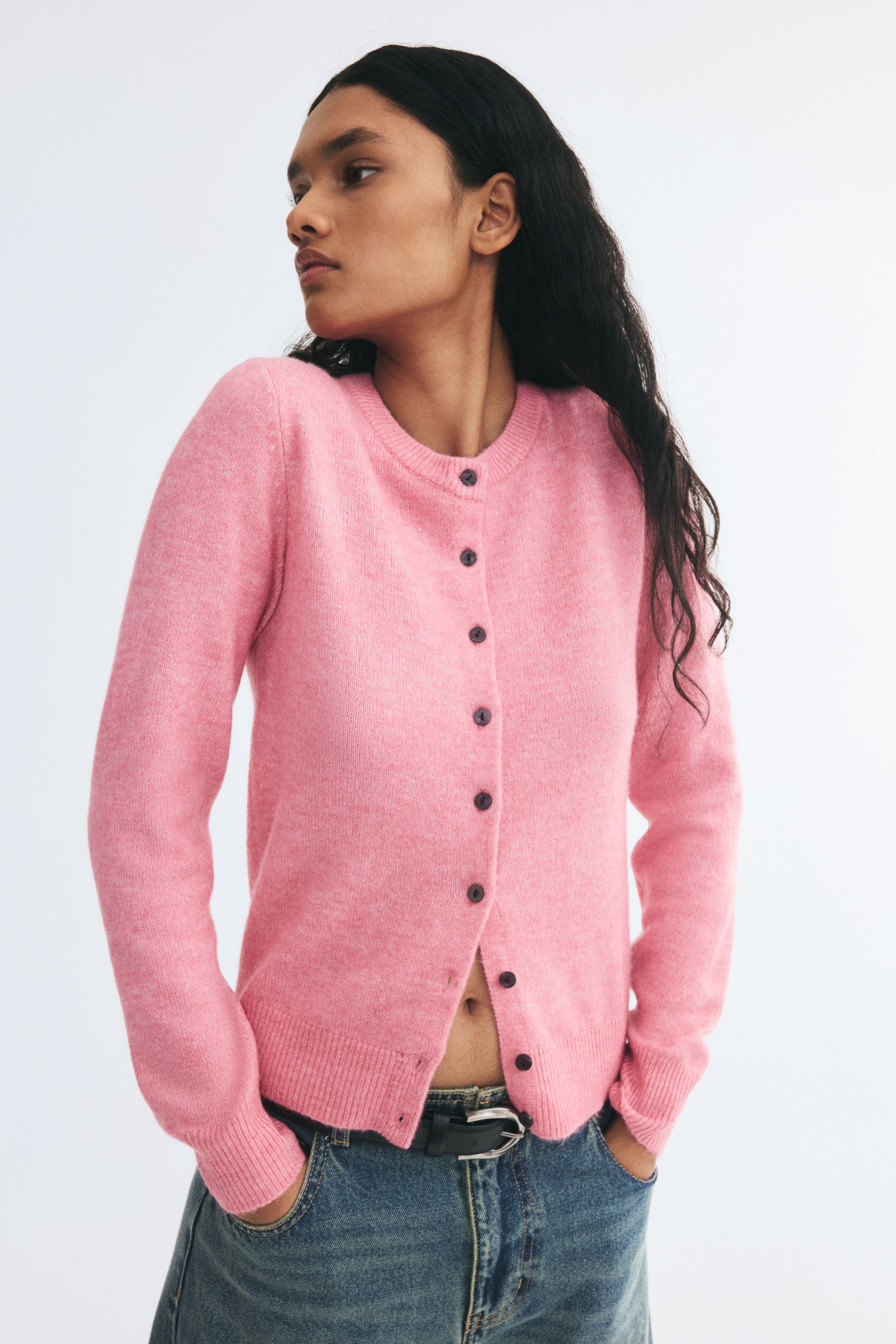 View larger image: A pink, fitted cardigan in a soft knit is worn by the model, featuring a ribbed crew neckline, dark front buttons, and ribbed cuffs and hem. The cardigan is styled with the bottom partially unbuttoned and paired with blue jeans and a black belt, while the model stands with hands in her pockets.