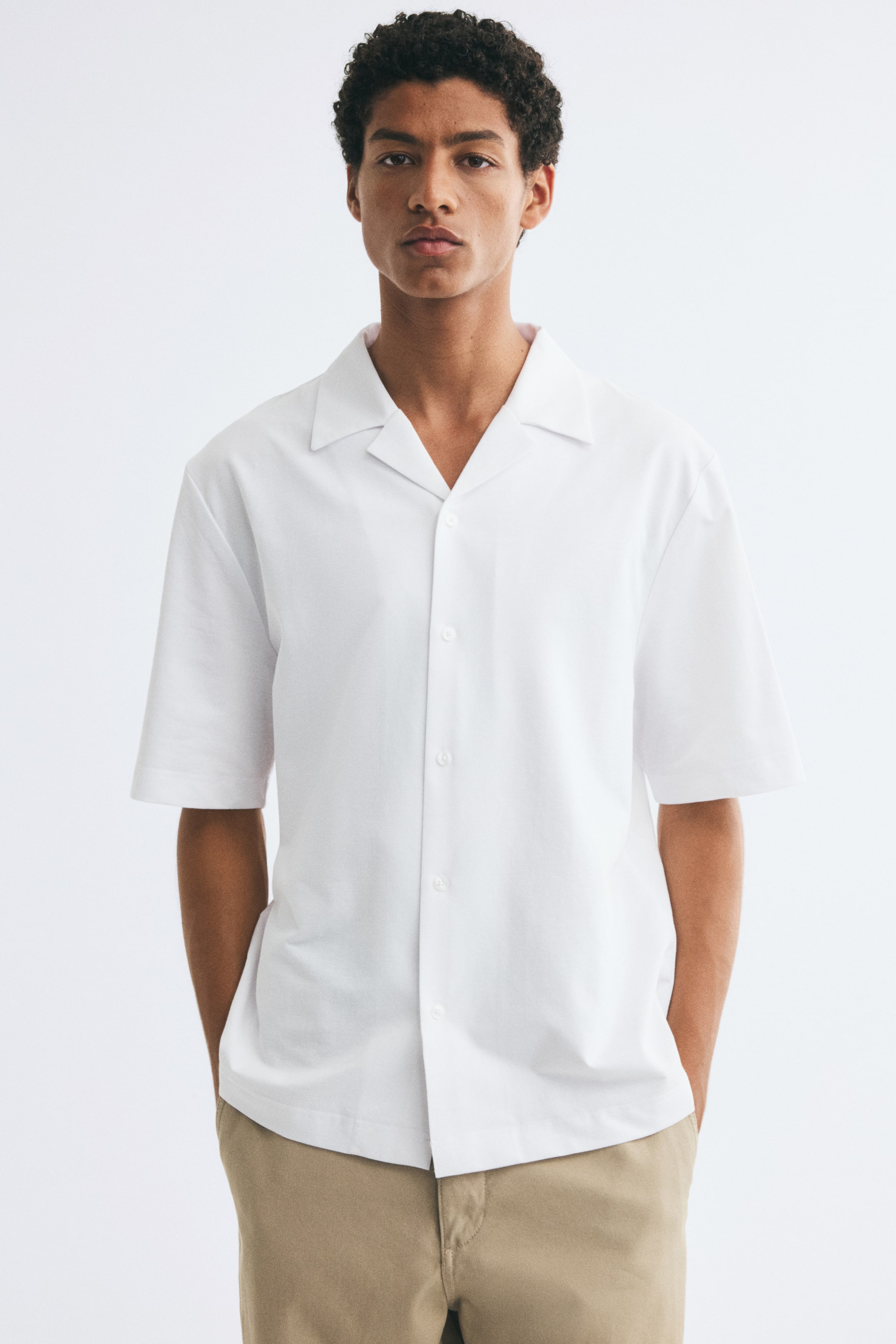 View larger image: The model is wearing a white short-sleeve shirt, styled untucked over beige trousers. The shirt features a resort collar, button-up front, and straight hem.