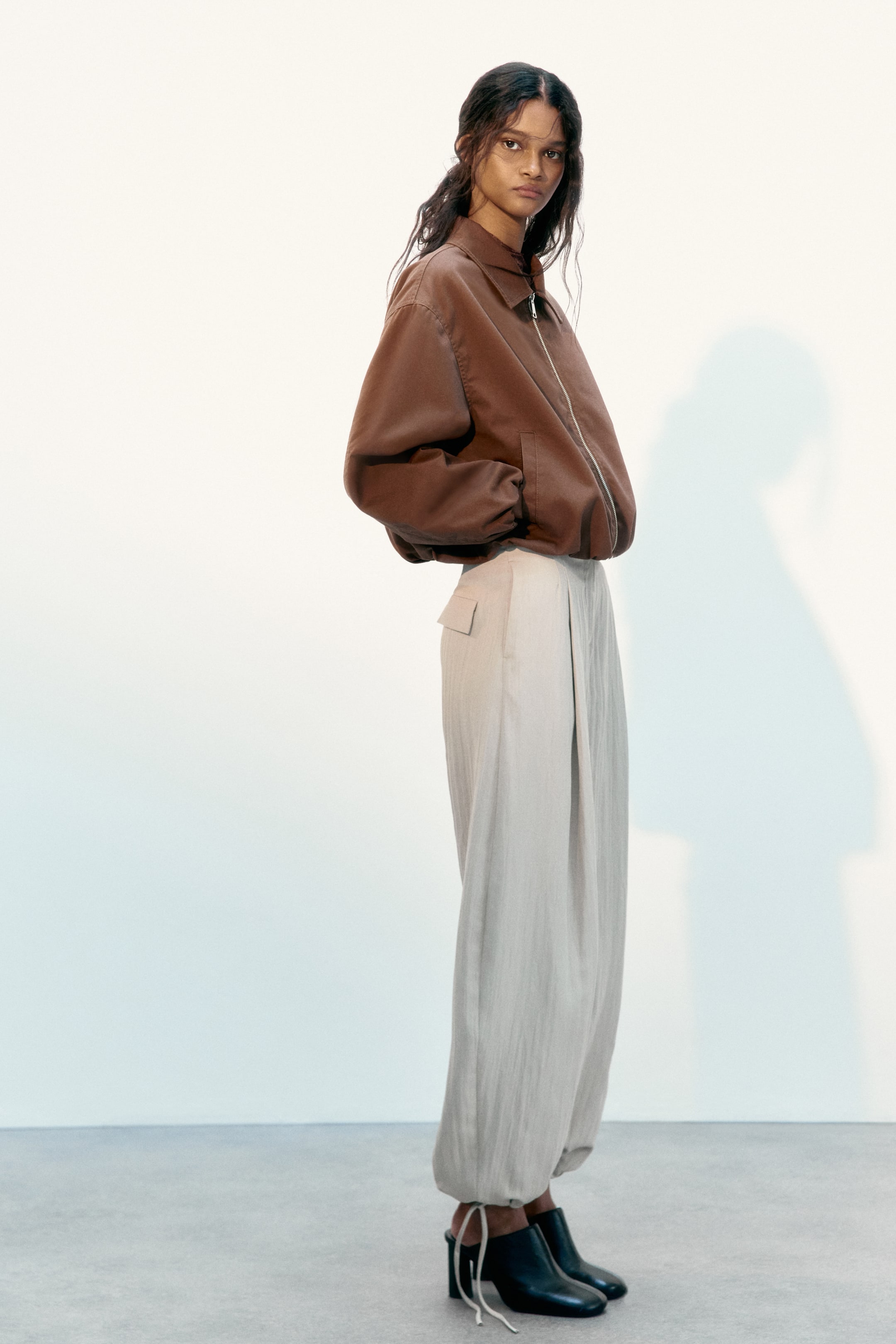 Vezi imaginea mai mare: Pantalonii largi de culoare bej deschis sunt confecționați dintr-un material texturat, cu mai multe pliuri în față, un buzunar cu clapă pe șoldul drept și glezne adunate cu șnururi vizibile, prezentând o siluetă relaxată și voluminoasă.