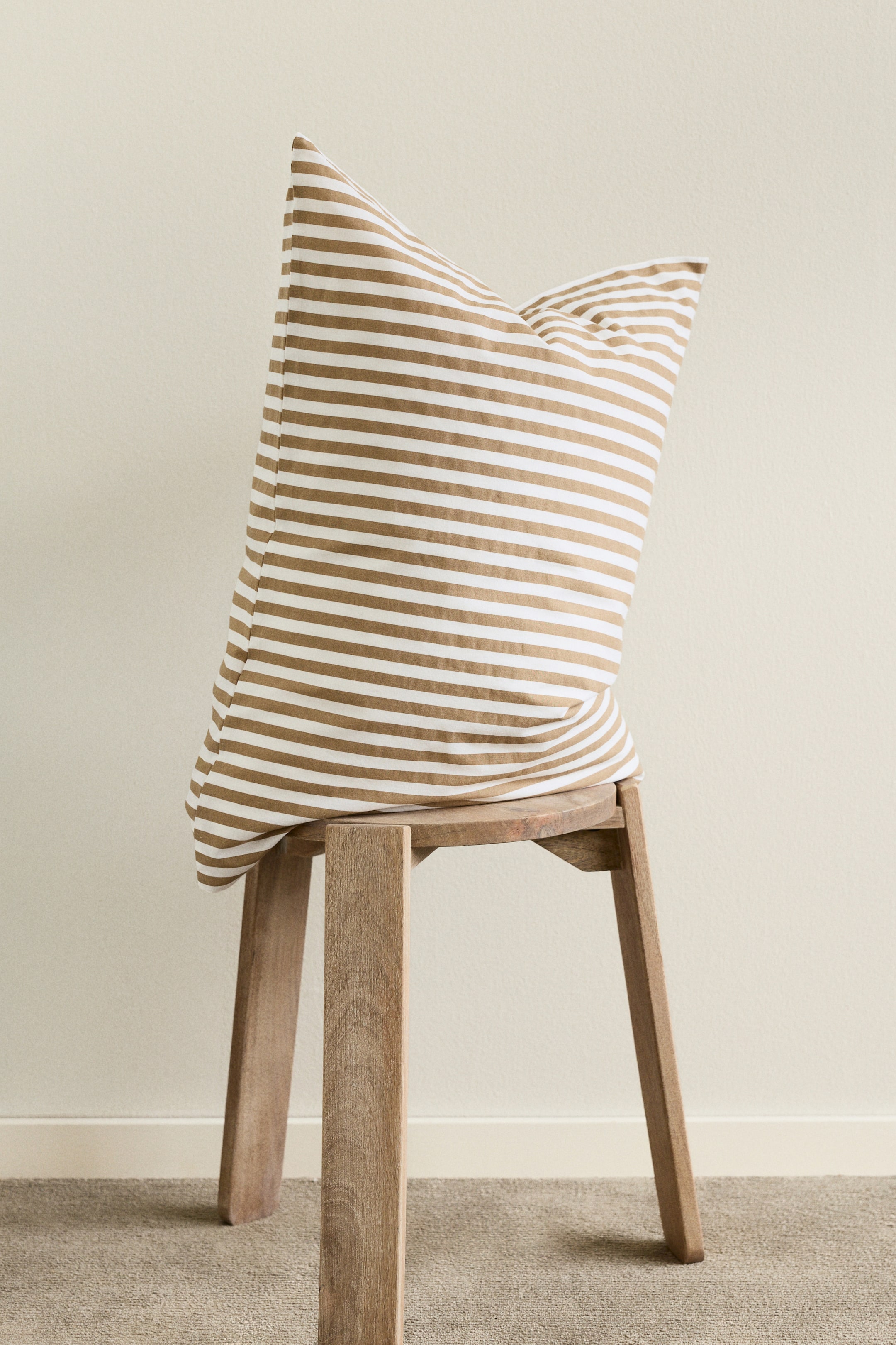 Se større billede: Et stribet pudebetræk vises i front, med skiftende vandrette striber i hvid og lysebrun. Det præsenterer en fyldig form, lavet af bomuldsvæv og inkluderer en kuvertlukning på bagsiden.