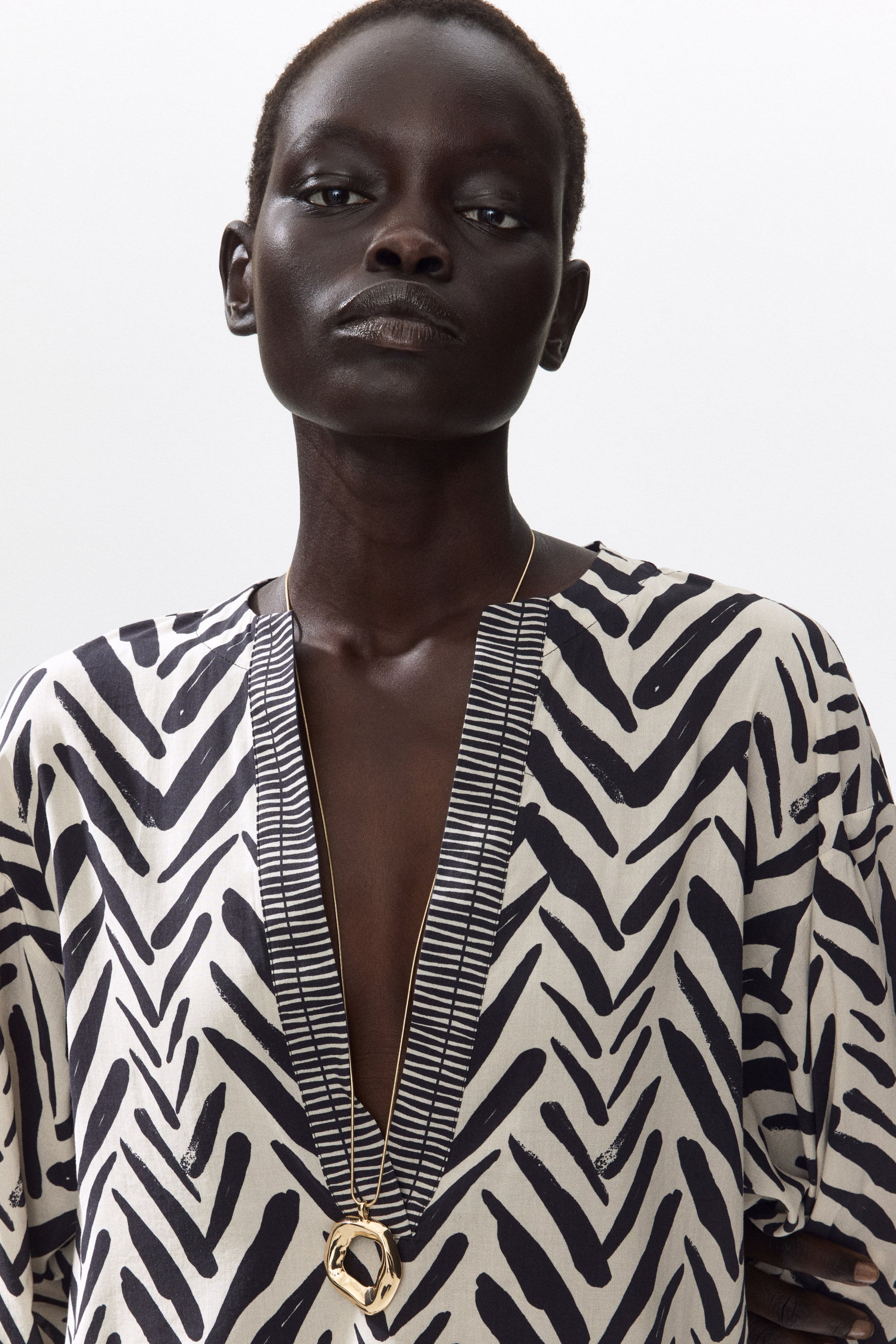 View larger image: The model is wearing a calf-length tunic dress adorned with a black and off-white abstract pattern. It showcases a deep V-neckline accentuated by a contrasting striped trim and features dropped shoulders. A gold-toned pendant necklace elegantly rests on the neckline.