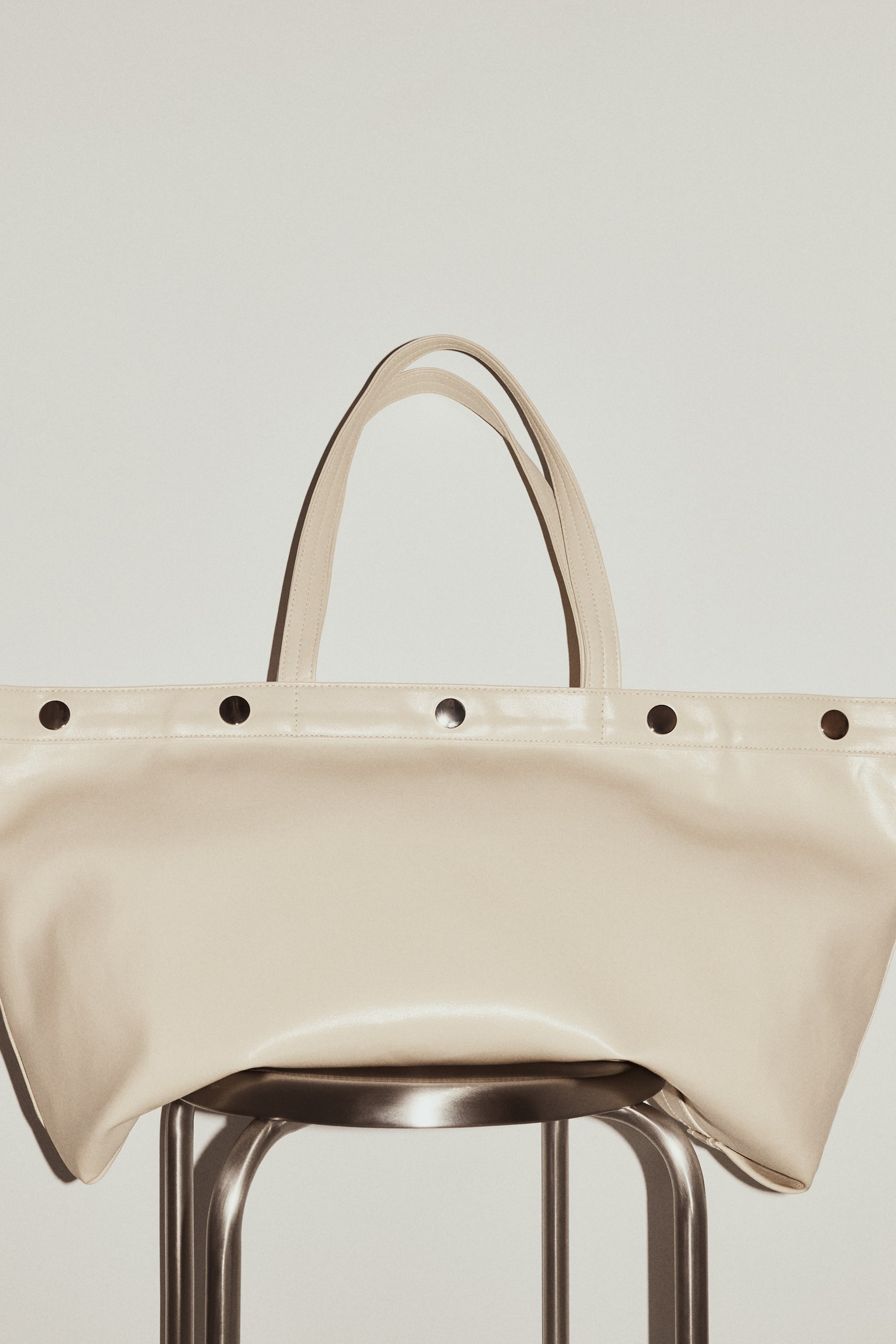 查看大圖: 奶油色托特包以正面朝外置於金屬凳上展示。包款配有兩條向上弧起的上提把，包口上緣飾有一列深色圓形鉚釘，柔軟材質則輕輕覆於凳面。
