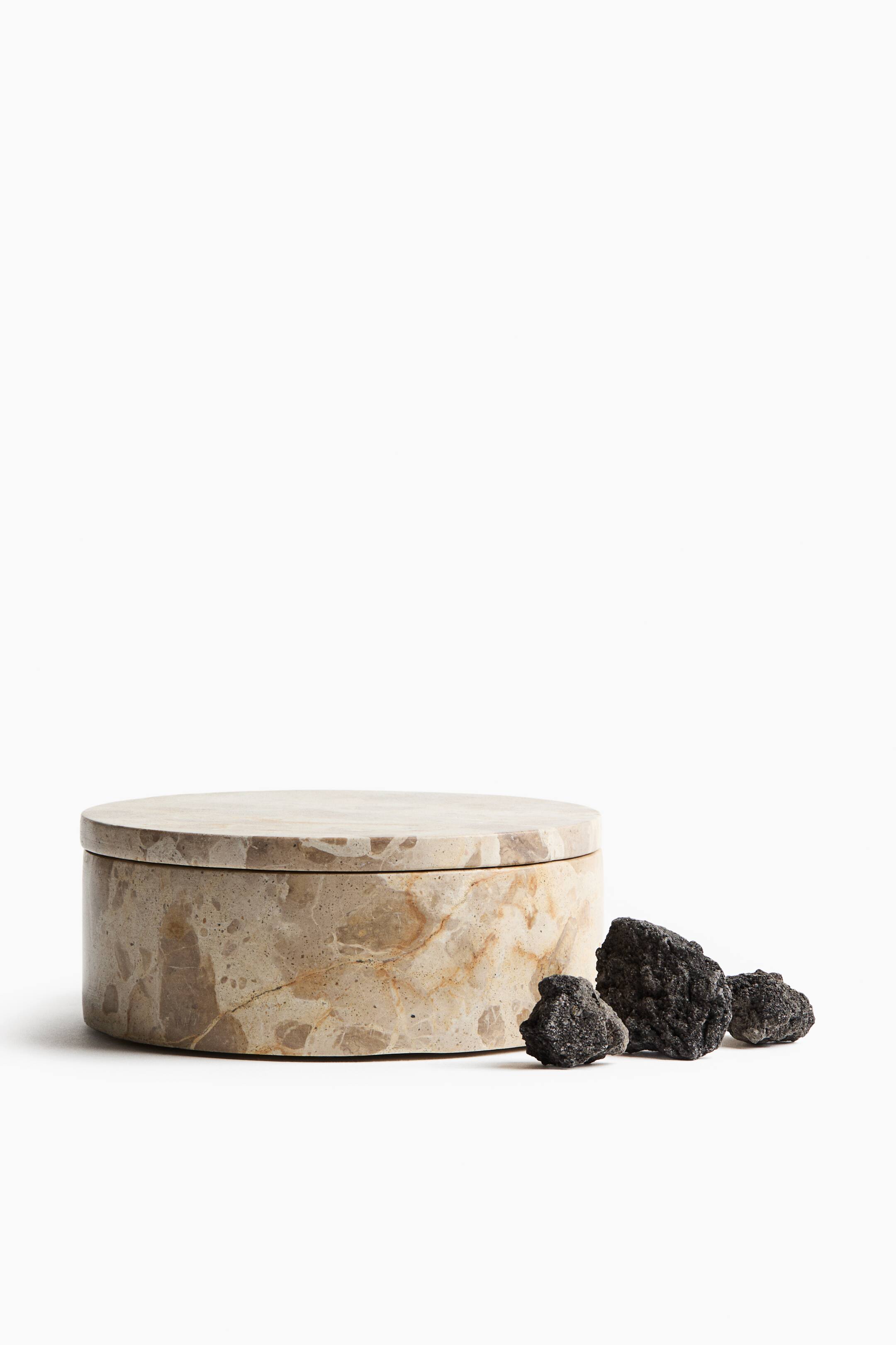 View larger image: A round beige marble container with a flat matching lid is positioned facing front, showing natural brown and tan veining. Three small textured black lava rocks are resting on the surface directly to its right.