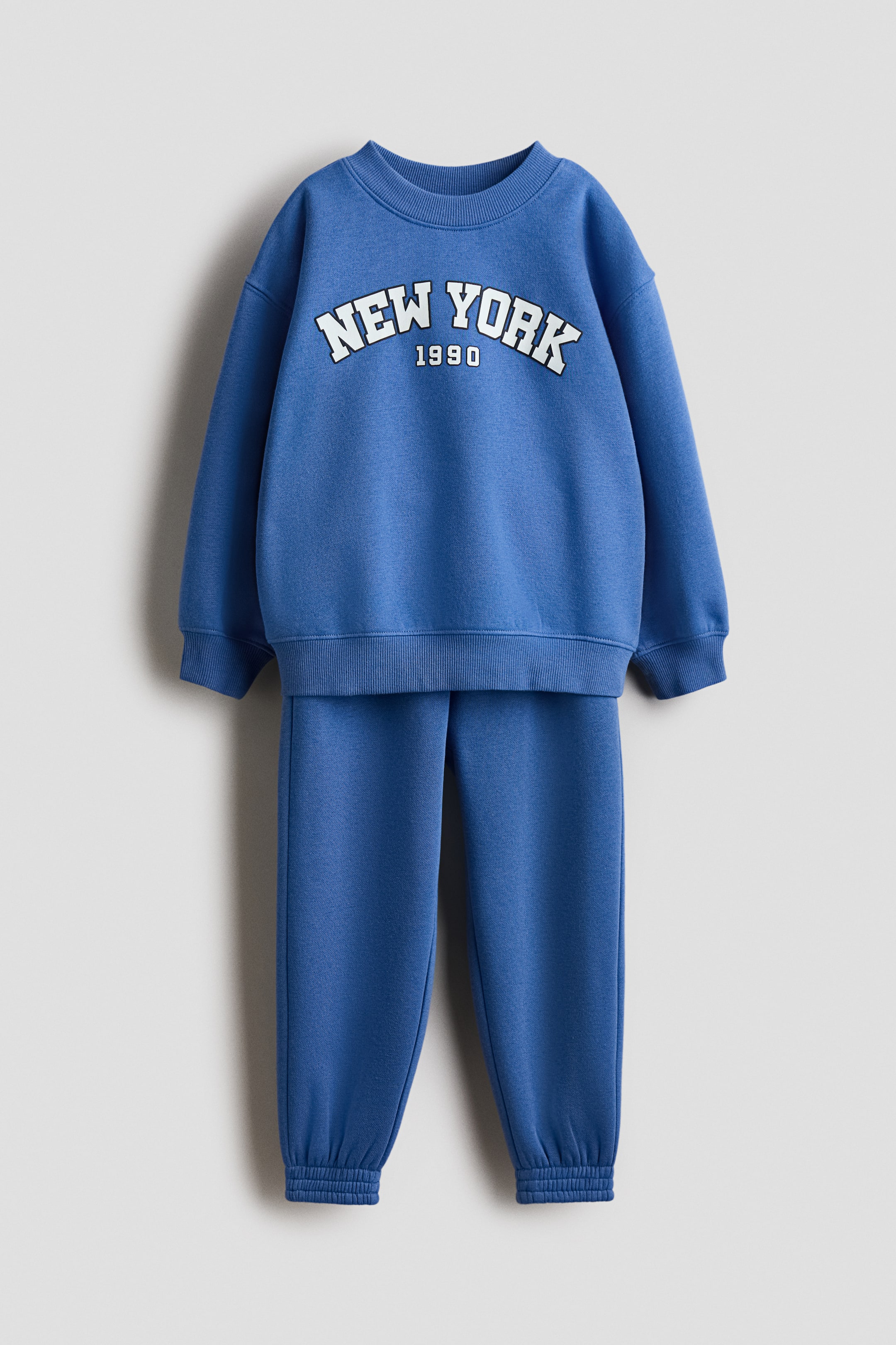 View larger image: A blue set, consisting of a lightweight sweatshirt and joggers, is presented in a FRONT view. The sweatshirt features a white "NEW YORK 1990" print across the chest, a round ribbed neckline, dropped shoulders, and ribbed cuffs and hem. The joggers have an elasticated waist and ribbed ankle cuffs.