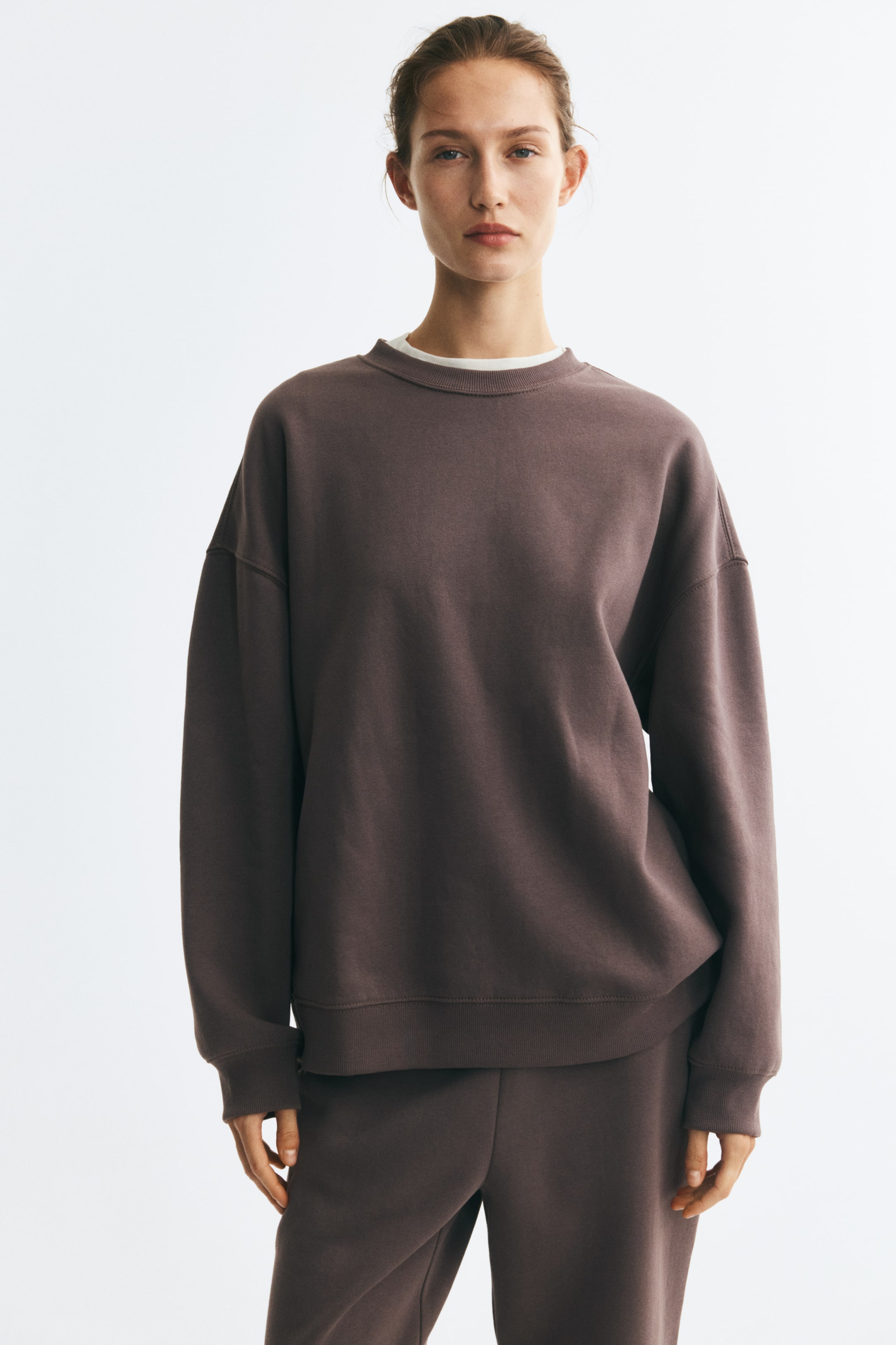 Grotere afbeelding bekijken: Een oversized bruine sweatshirt met ronde hals, afhangende schouders en geribde boorden wordt gedragen door het model. De sweatshirt is gelaagd over een zichtbaar wit kraagje en gecombineerd met een bijpassende bruine joggingbroek.