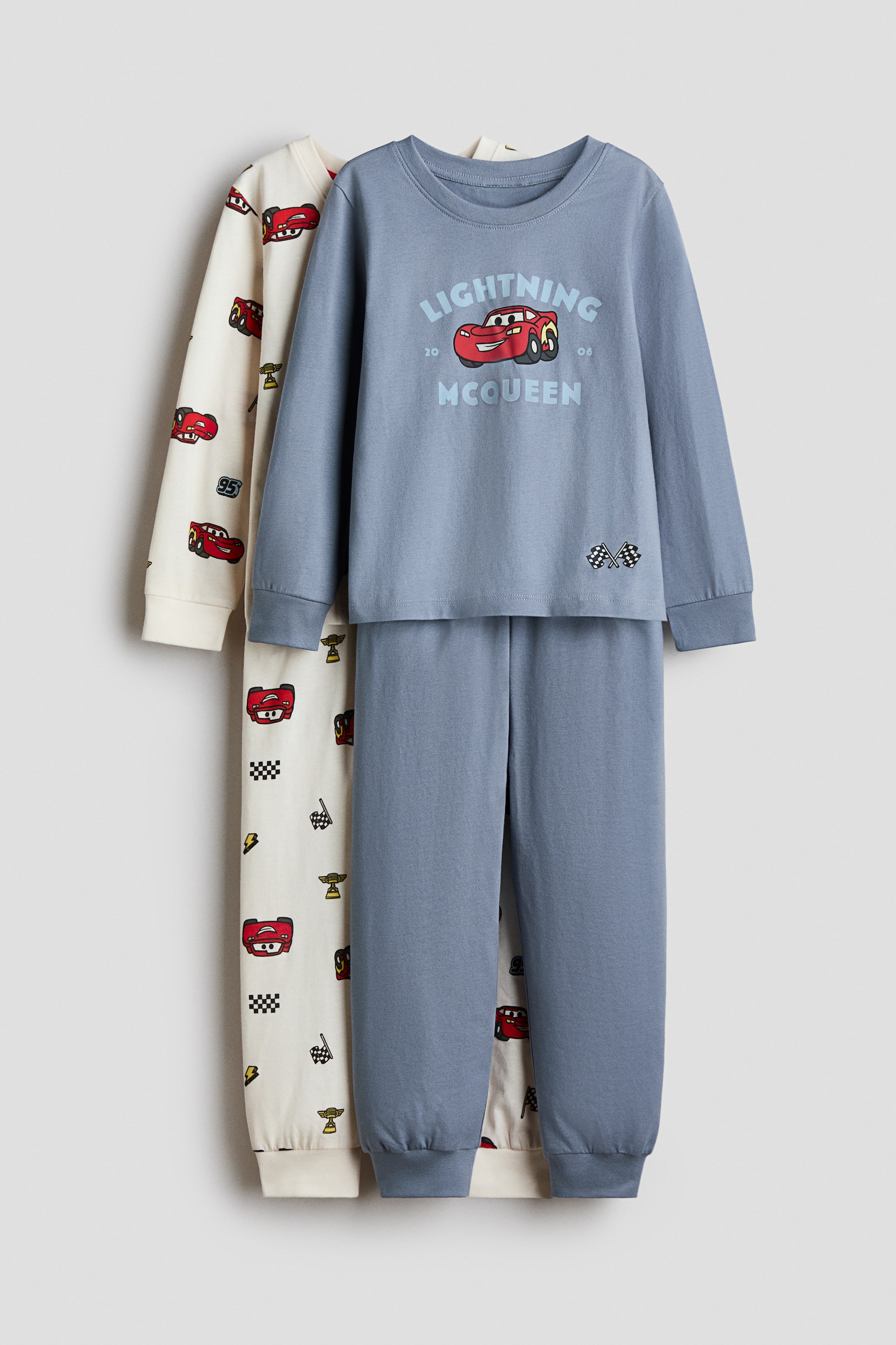 View larger image: Two pyjama top and pants sets are hanging facing front, with a dusty blue set layered over an off-white set. The blue set features a long-sleeve top with a central "Lightning McQueen" graphic and ribbed cuffs, paired with matching pants with ribbed hems. The off-white set displays an all-over print of red racecars, checkered flags, and trophies on both the long-sleeve top and pants.