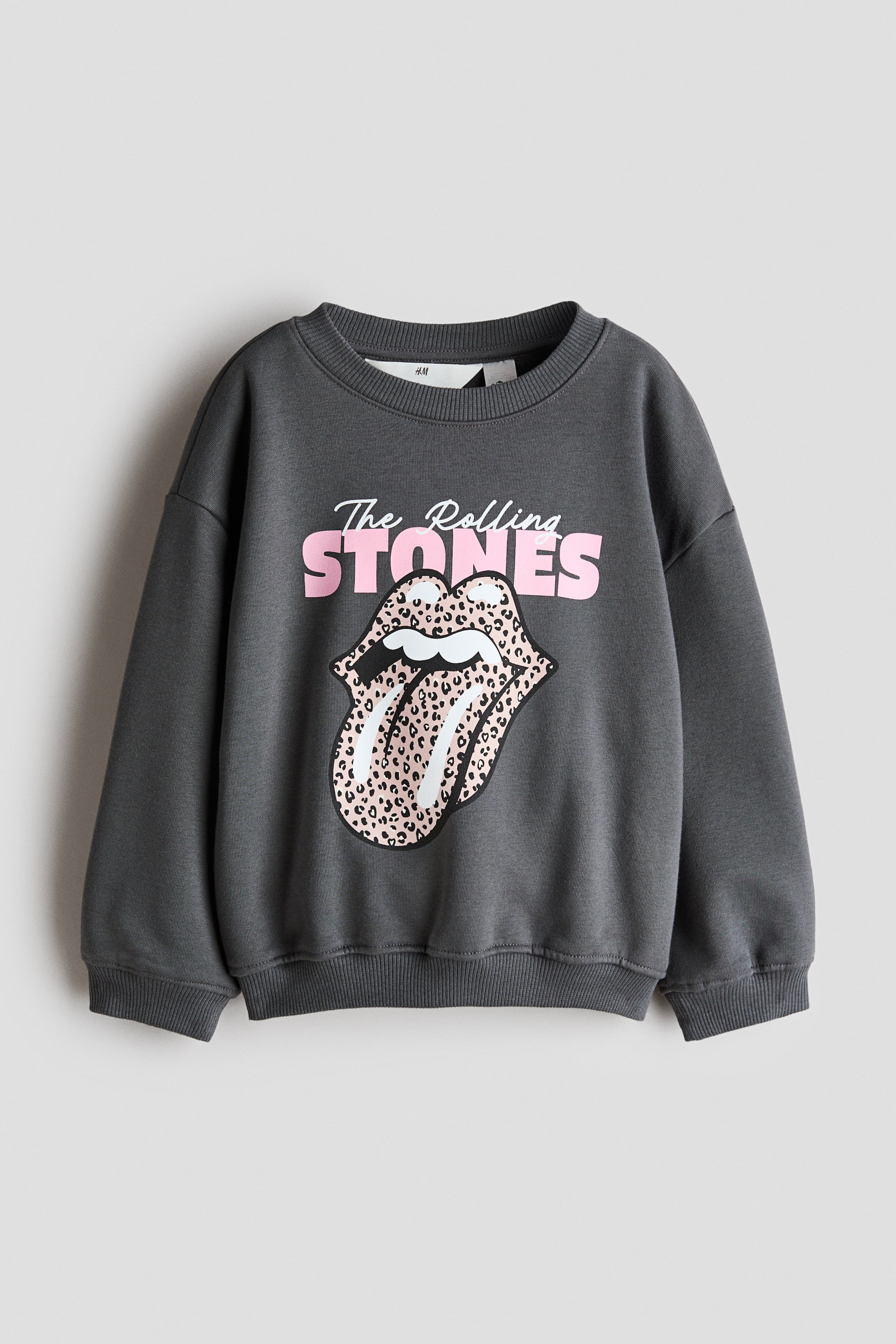 Agrandir l'image: Un sweat-shirt oversize gris foncé est présenté de face, avec un col rond côtelé, des épaules tombantes, ainsi que des poignets et un ourlet côtelés. L’avant arbore un imprimé graphique « The Rolling Stones » en lettrage rose et blanc, au-dessus d’un logo langue et lèvres à motif léopard rose.