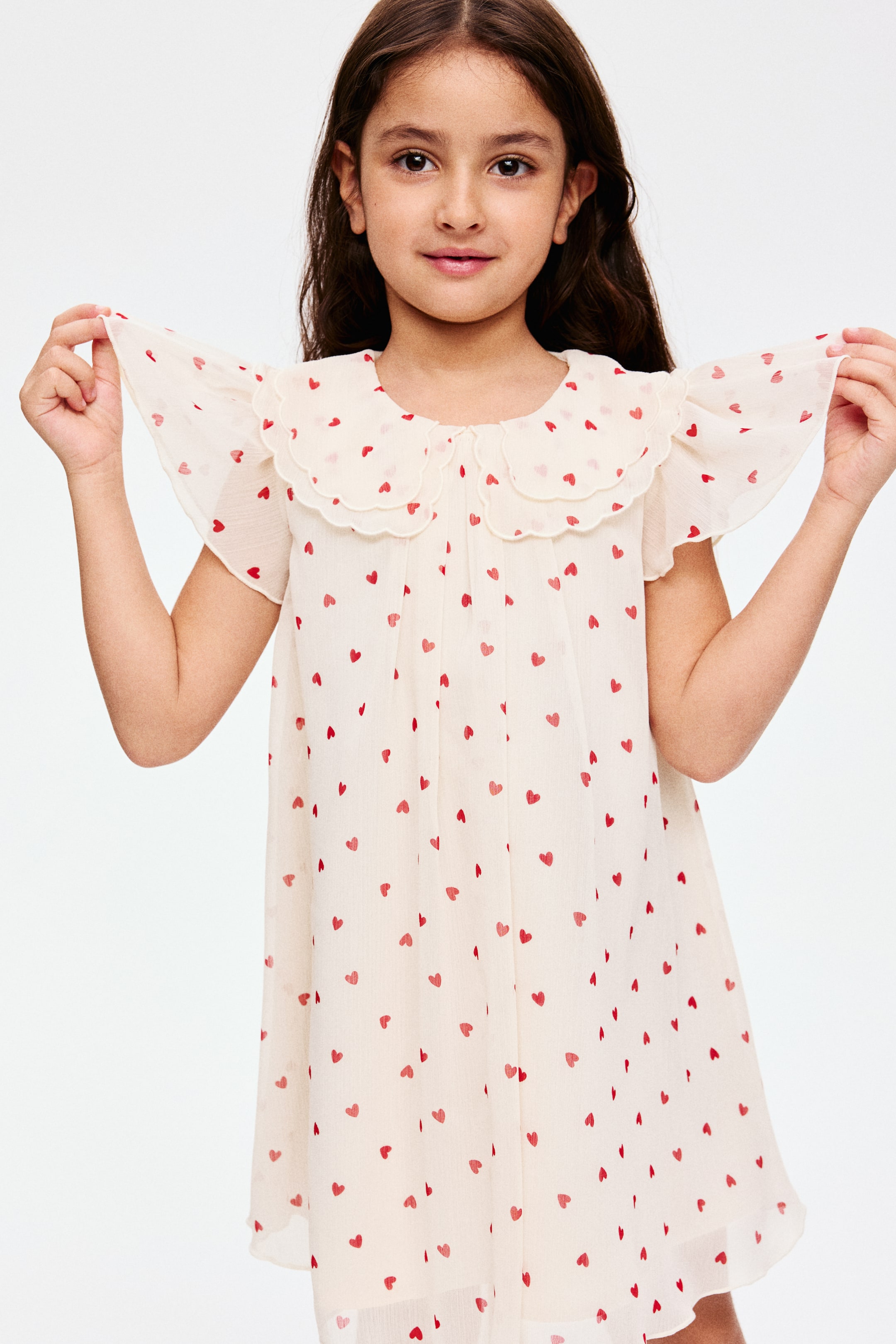 Prikaži veću sliku: Haljina A-kroja, prikazana s PREDNJE strane, odlikuje se svetlom šifonom sa uzorkom crvenih srca, okruglim izrezom, dvostrukim slojevitim Peter Pan kragnom talasastih ivica, kratkim leptir rukavima i ravnim rubom.