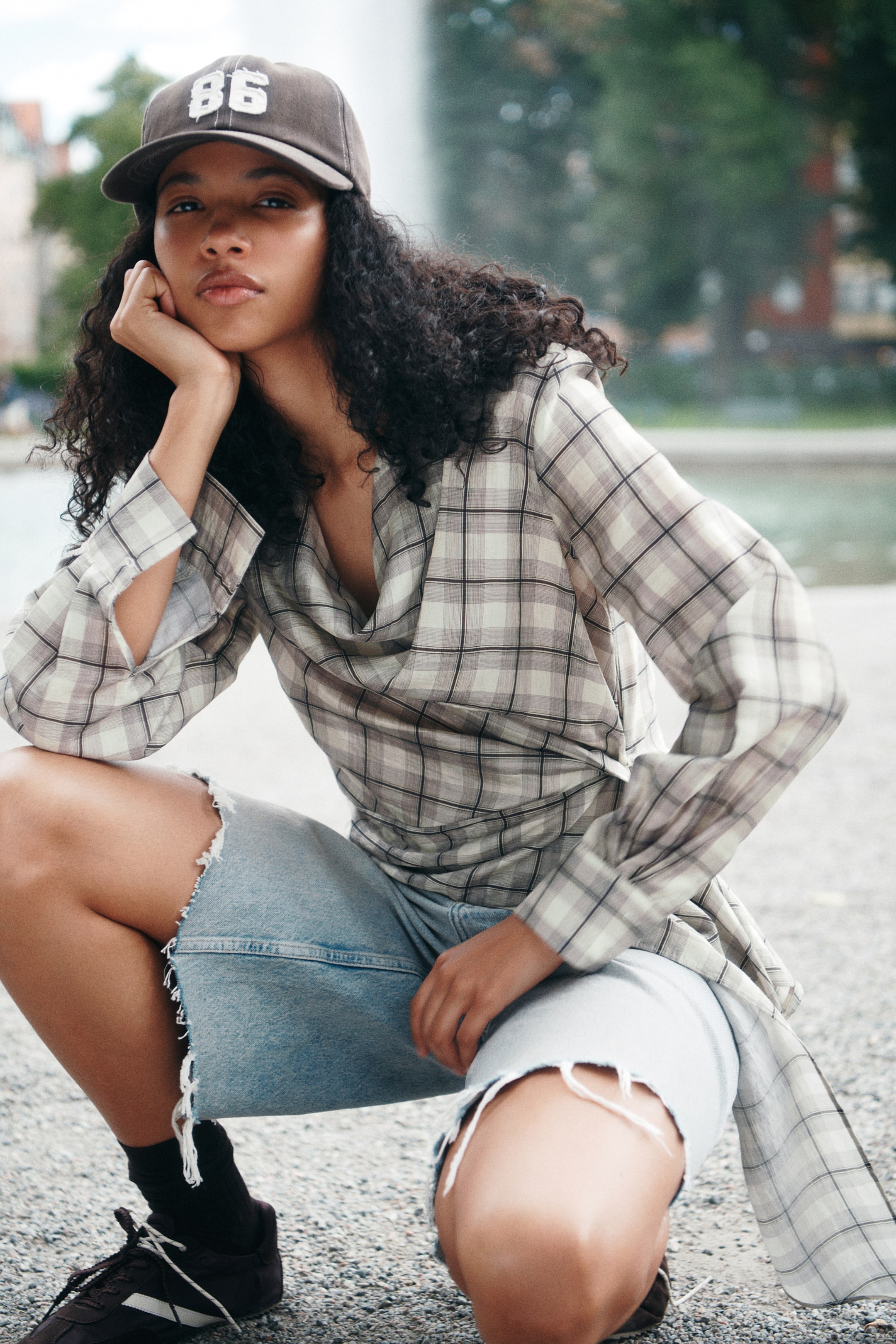 Ampliar la imagen: La modelo, en posición de cuclillas, lleva una blusa de cuadros con escote drapeado, frente con caída y un panel lateral largo y colgante. La blusa de manga larga se combina con shorts de mezclilla de lavado claro y deshilachados, así como con una gorra de béisbol café.