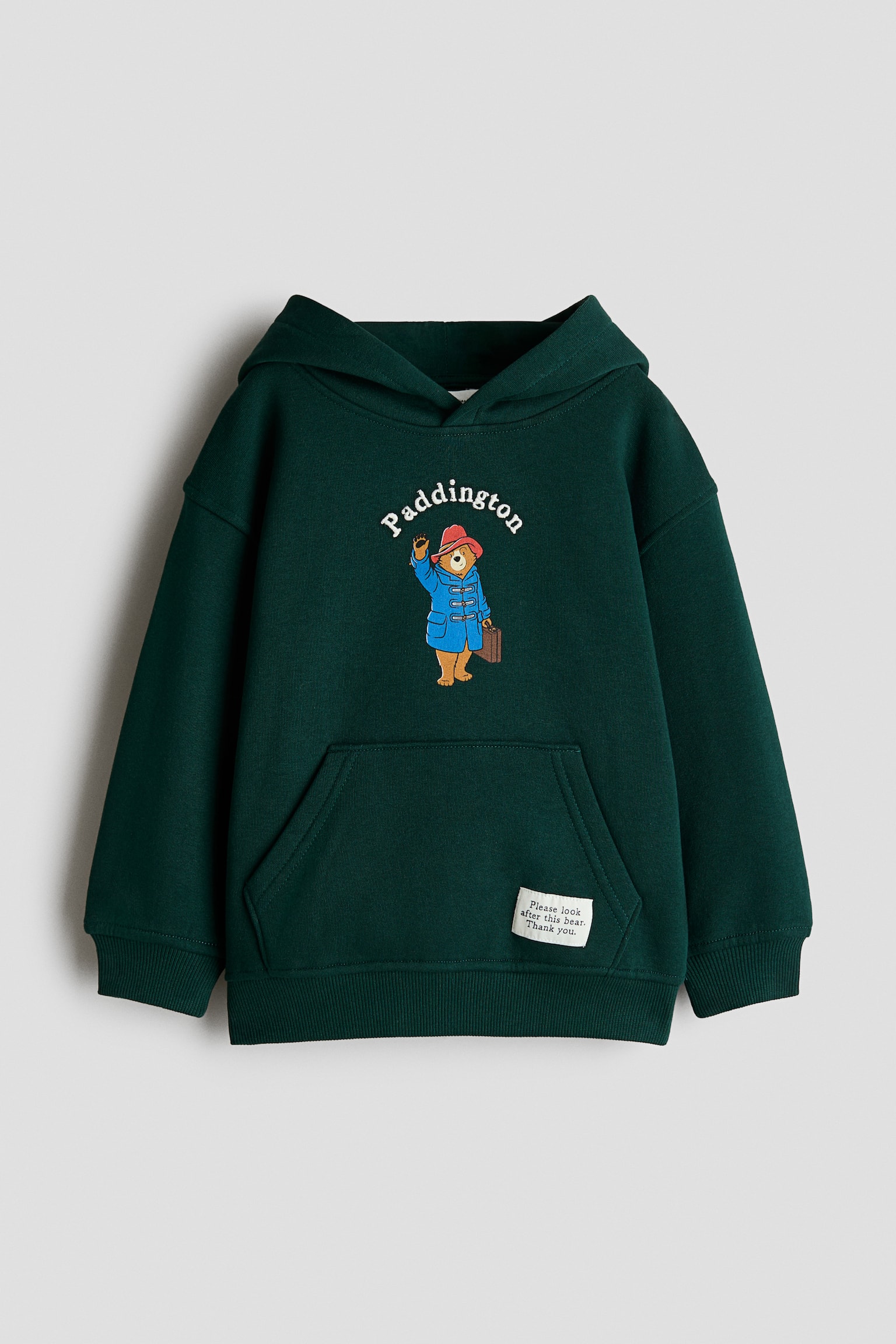 Een donkergroene hoodie met losse pasvorm wordt van voren getoond en is voorzien van een omslaande capuchon, verlaagde schouders, een kangoeroezak en ribgebreide boorden en zoom. Op de borst staat een geprint Paddington Bear-illustratie met gebogen witte tekst erboven, samen met een klein wit label op de voorzak.