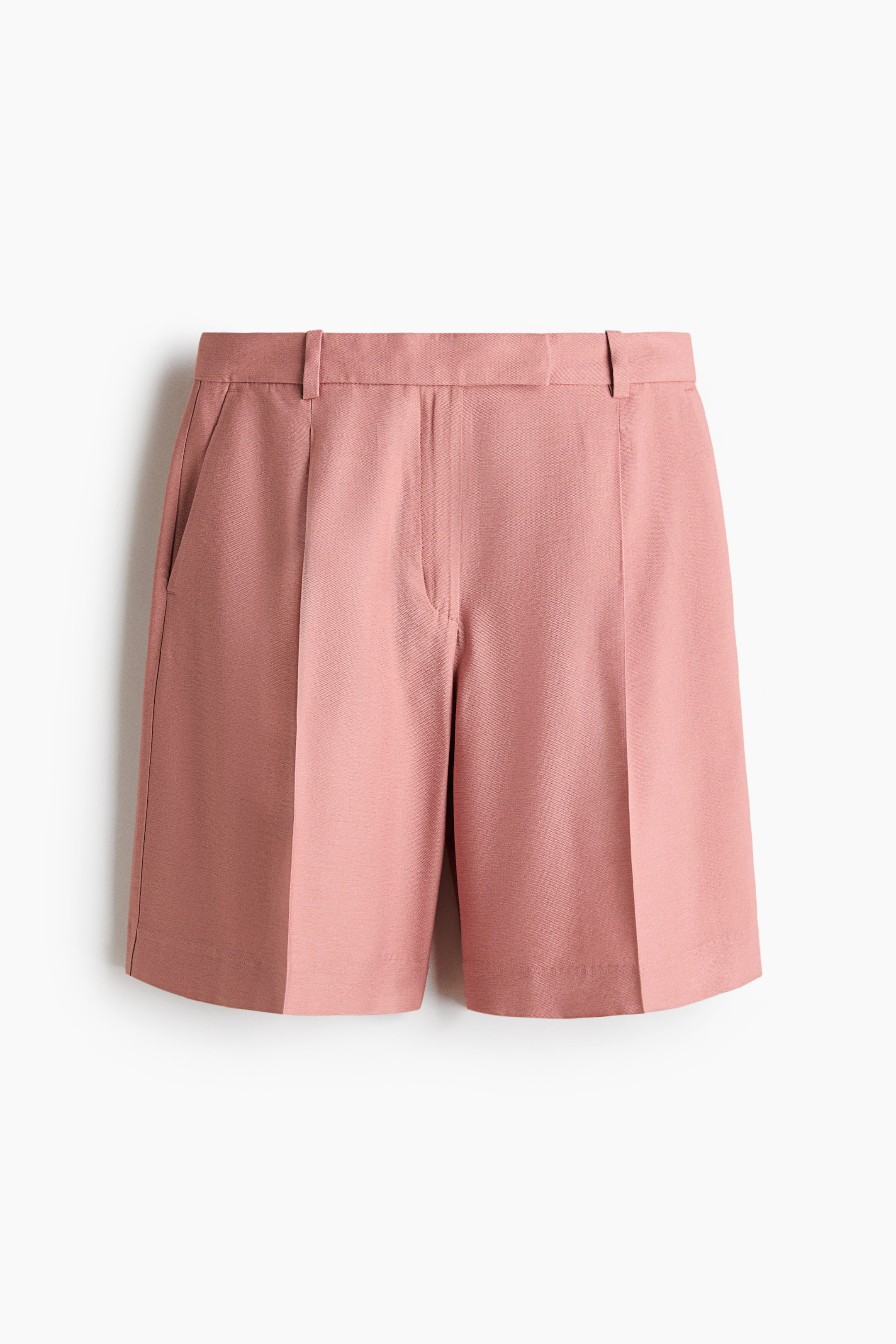 View larger image: The pink knee-length, loose-fit shorts are shown from the front, featuring an extended waistband with visible belt loops and a prominent zip fly. Diagonal side pockets are visible, with pleats and creases running down the front of the legs.