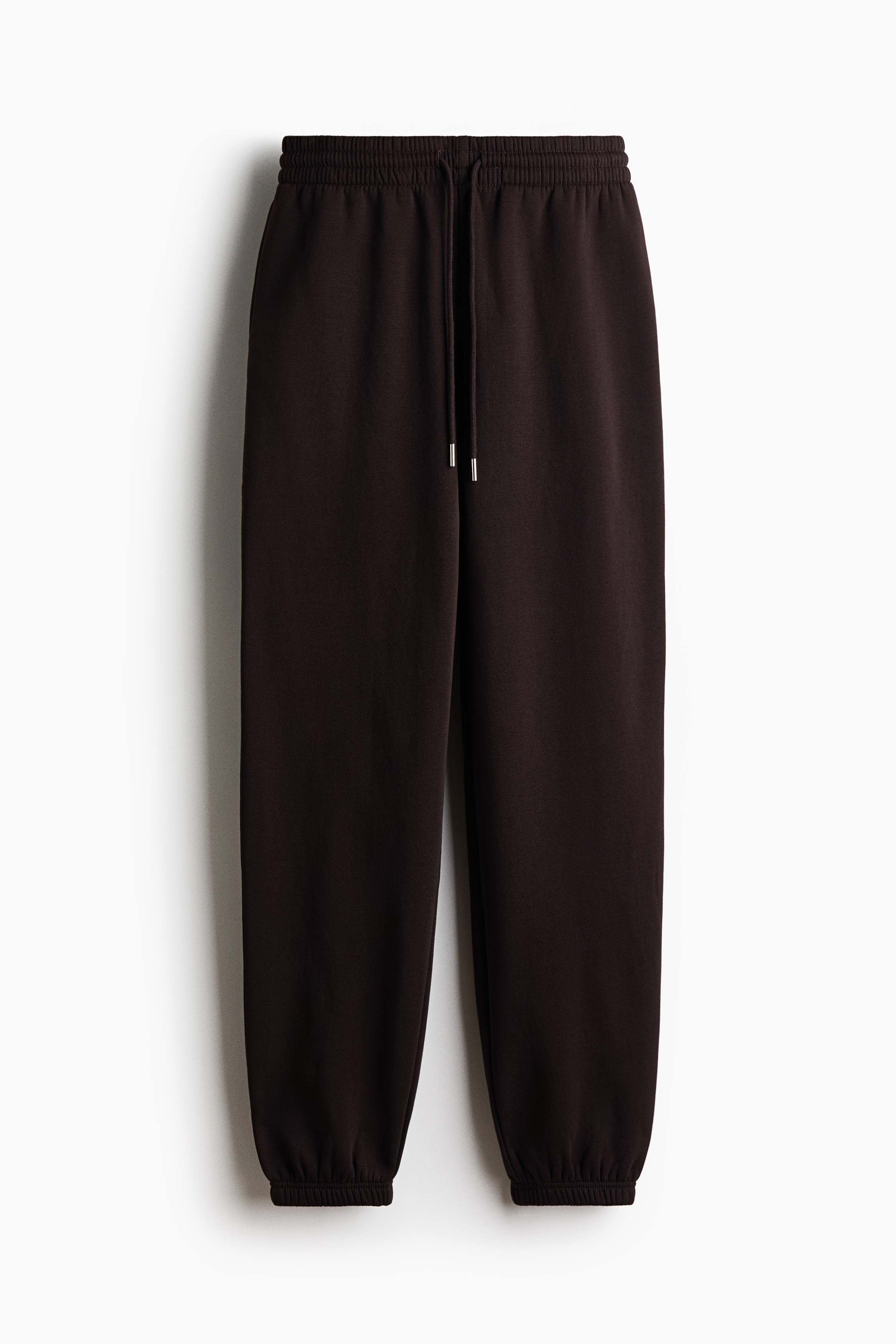 Track pants in a dark brown sweat fabric are being worn by the model, who is standing in profile with one hand resting inside the side pocket. The high-waisted pants feature an elasticated waistband with a drawstring and gathered elastication at the hems, styled with a grey tank top and dark sneakers.