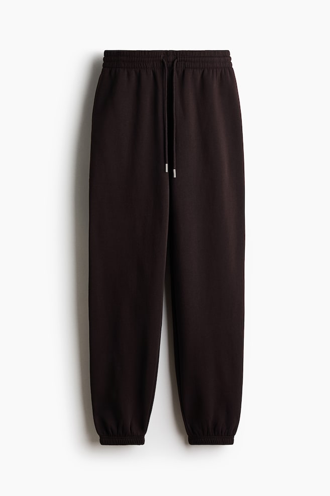 A pair of dark brown track pants is shown from the front, featuring a high, covered elastic waistband with a matching drawstring and tapering down to gathered elastic hems. - 1