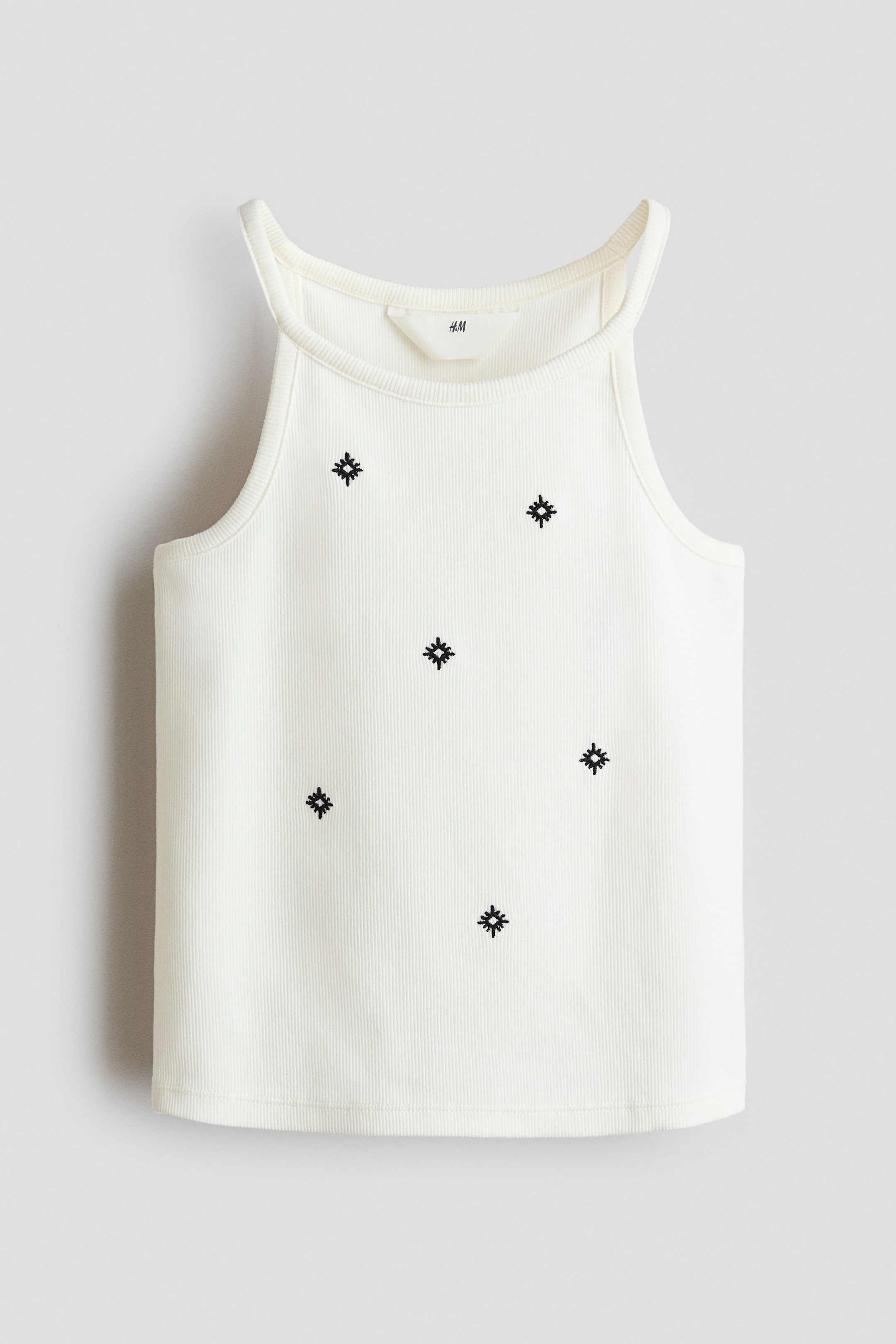 View larger image: Presenting the front view, this off-white ribbed tank top showcases a high, rounded neckline and slender shoulder straps. Adorning its vertically ribbed fabric are small black embroidered starburst patterns, adding a touch of elegance.