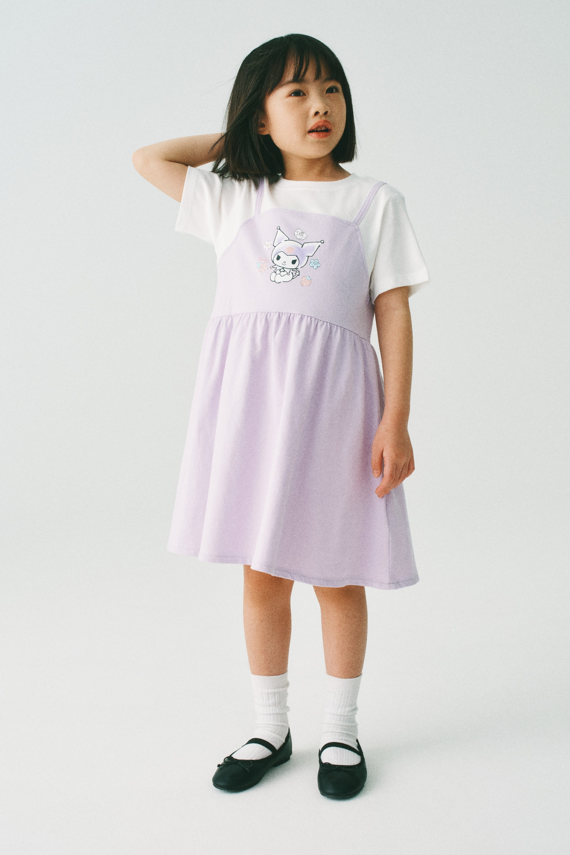 View larger image: The model is donning a layered dress. The lavender top layer showcases a square neckline, narrow shoulder straps, a gathered waist seam, and a character print motif on the front. Beneath, a sewn-in white underlayer is visible, featuring a round neckline and short sleeves.