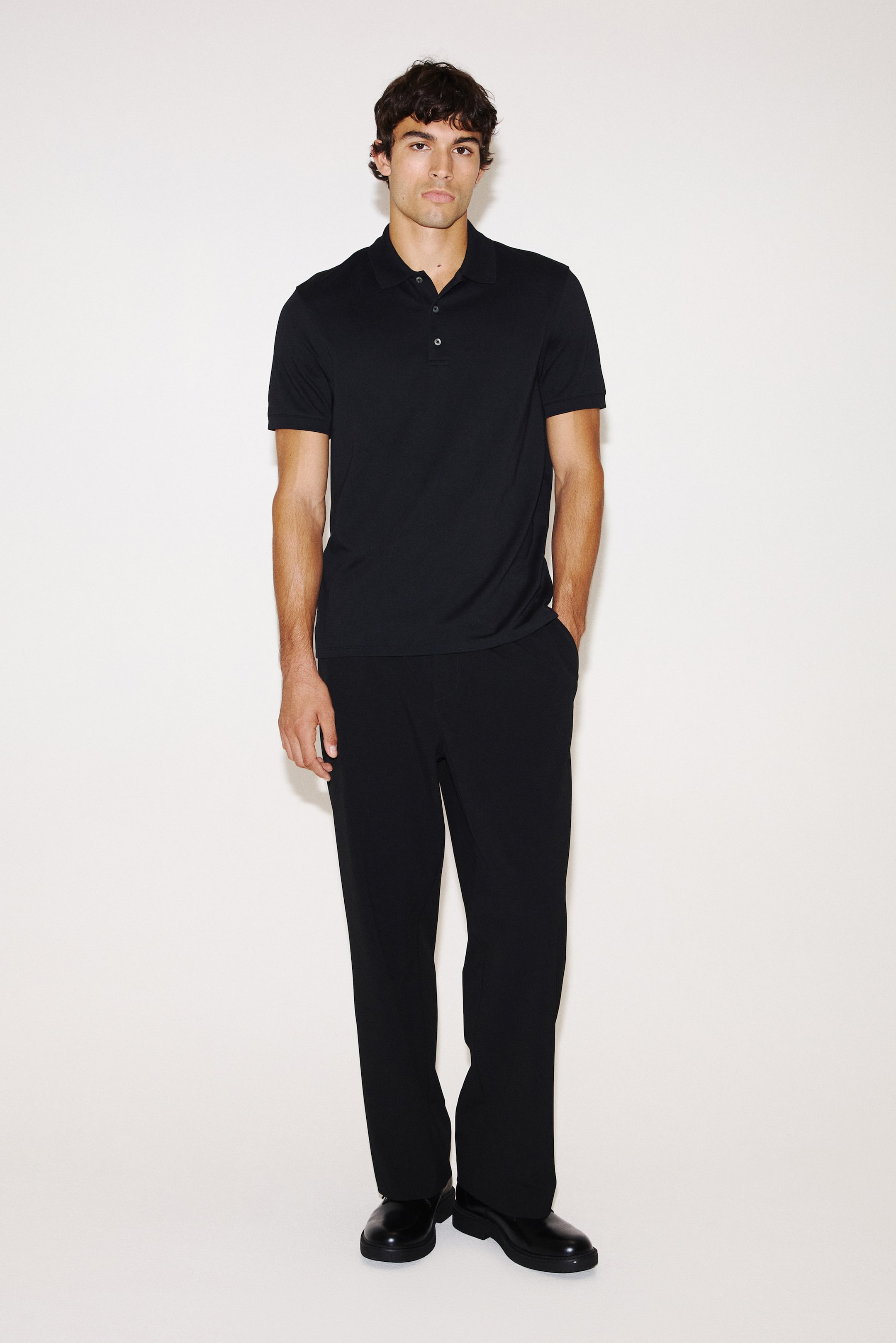 Ampliar la imagen: La modelo lleva puesta una playera polo negra con cuello clásico, tapeta de botones y mangas cortas con puños ribeteados. La playera de corte regular está combinada con pantalones negros a juego y zapatos negros.
