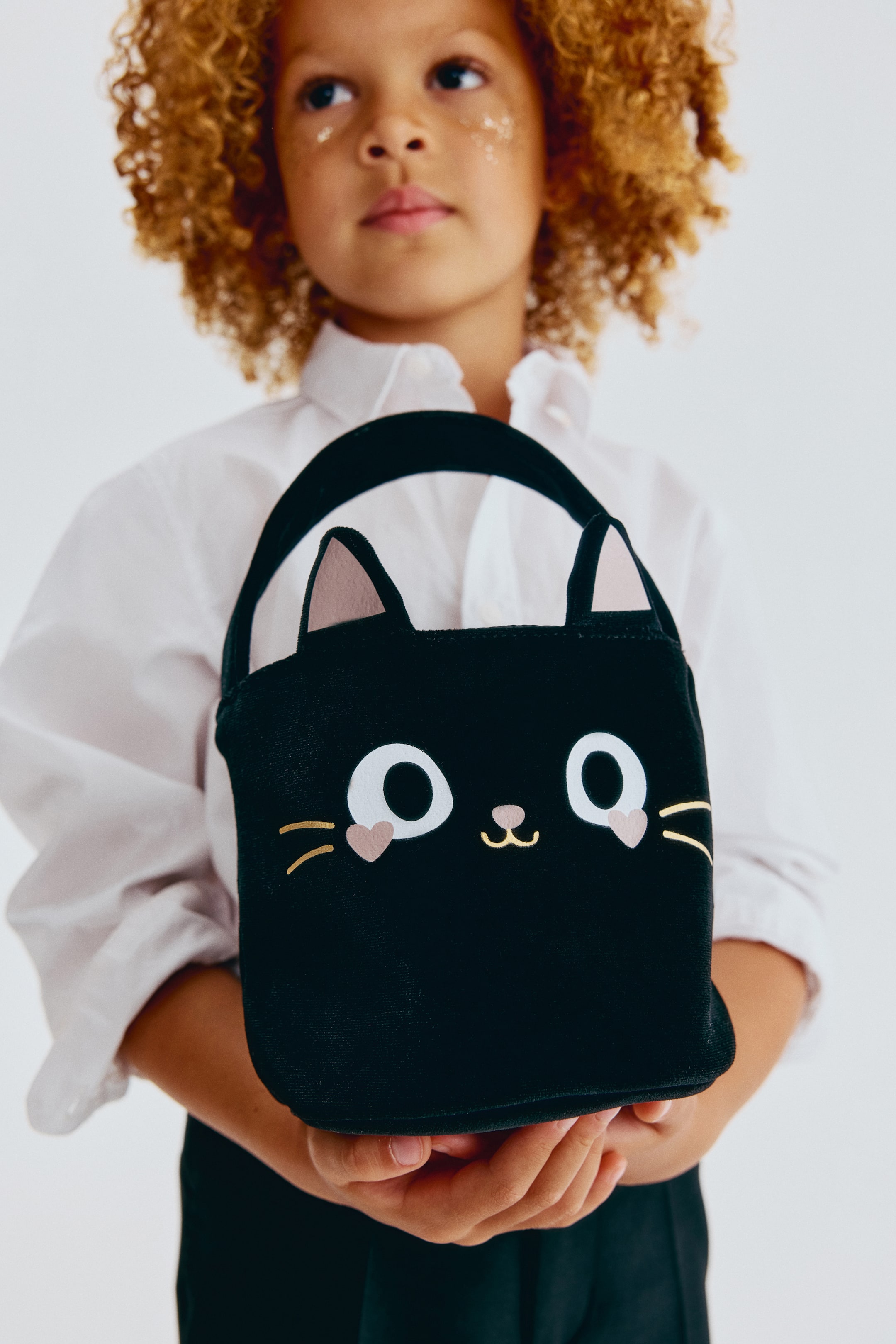 View larger image: The black cat-shaped bag is front-facing, featuring two pointed ears with light pink inner sections and a matching black handle. Its front panel displays large white circular eyes with small pink heart appliqués below them, a light pink nose appliqué, and gold embroidered whiskers and a curved mouth.