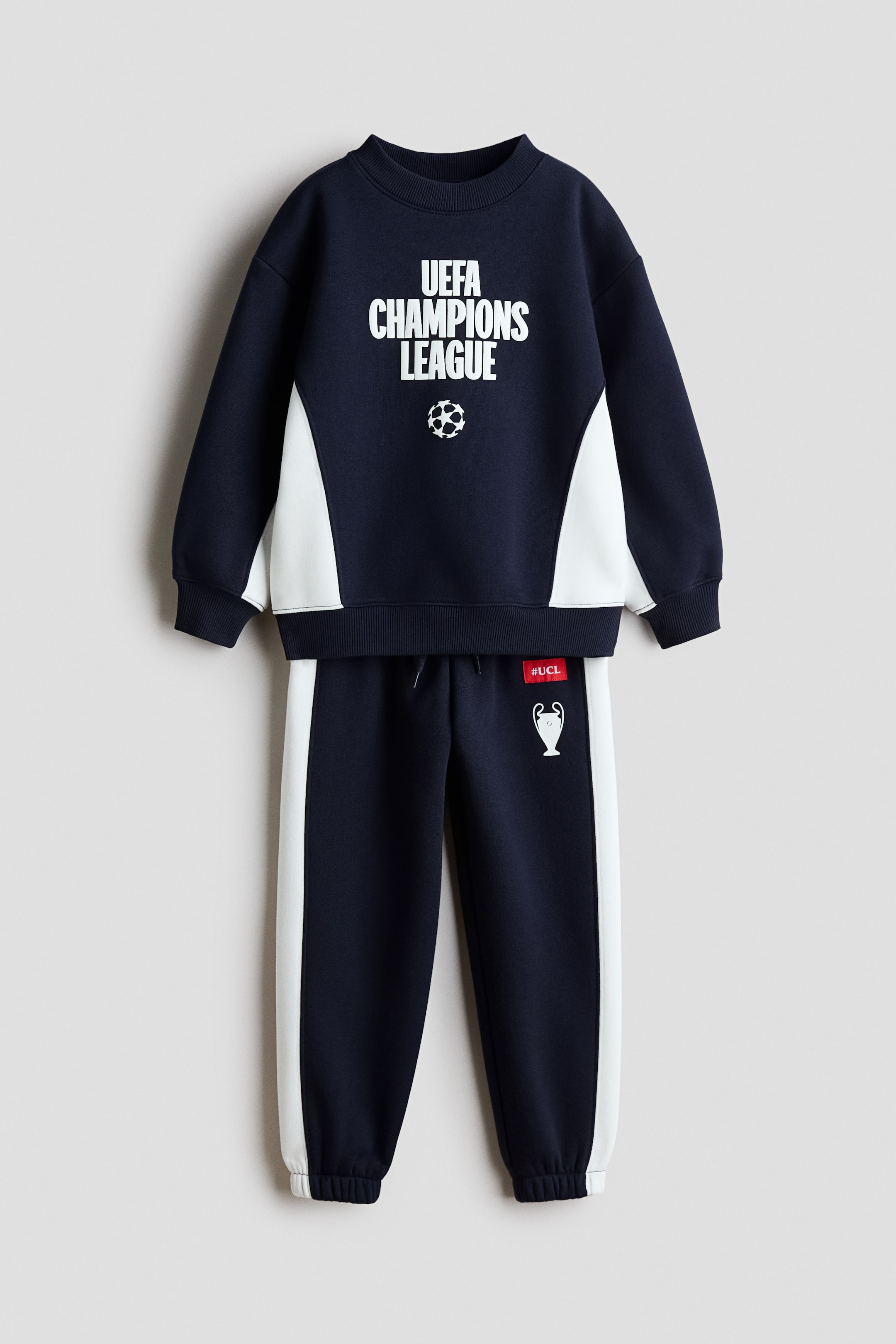 View larger image: A front-facing navy blue and white fleece sweatshirt and tracksuit bottoms set is displaying a printed sports design. The sweatshirt features a round, rib-trimmed neckline, ribbed cuffs and hem, and a "UEFA CHAMPIONS LEAGUE" graphic on the chest, while the matching tracksuit bottoms show white side stripes, an elasticated waist, and elasticated hems.