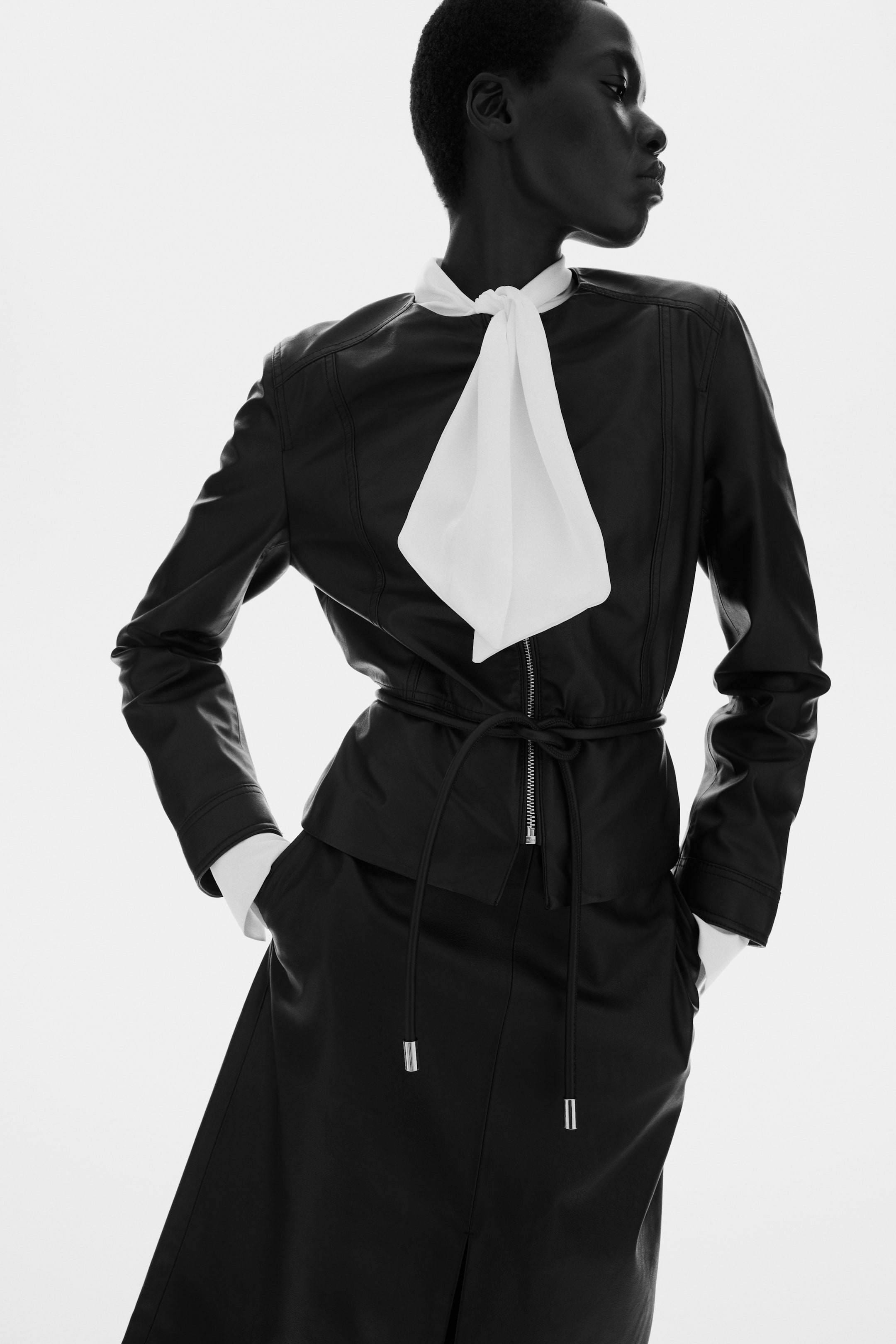 View larger image: The model is wearing a short, fitted jacket made of coated fabric, showcasing a round neckline, a partially unzipped front, and a tubular tie belt that cinches the waist above a flared peplum. Underneath the jacket, a white shirt with a large bow is layered, with its cuffs visibly extending.