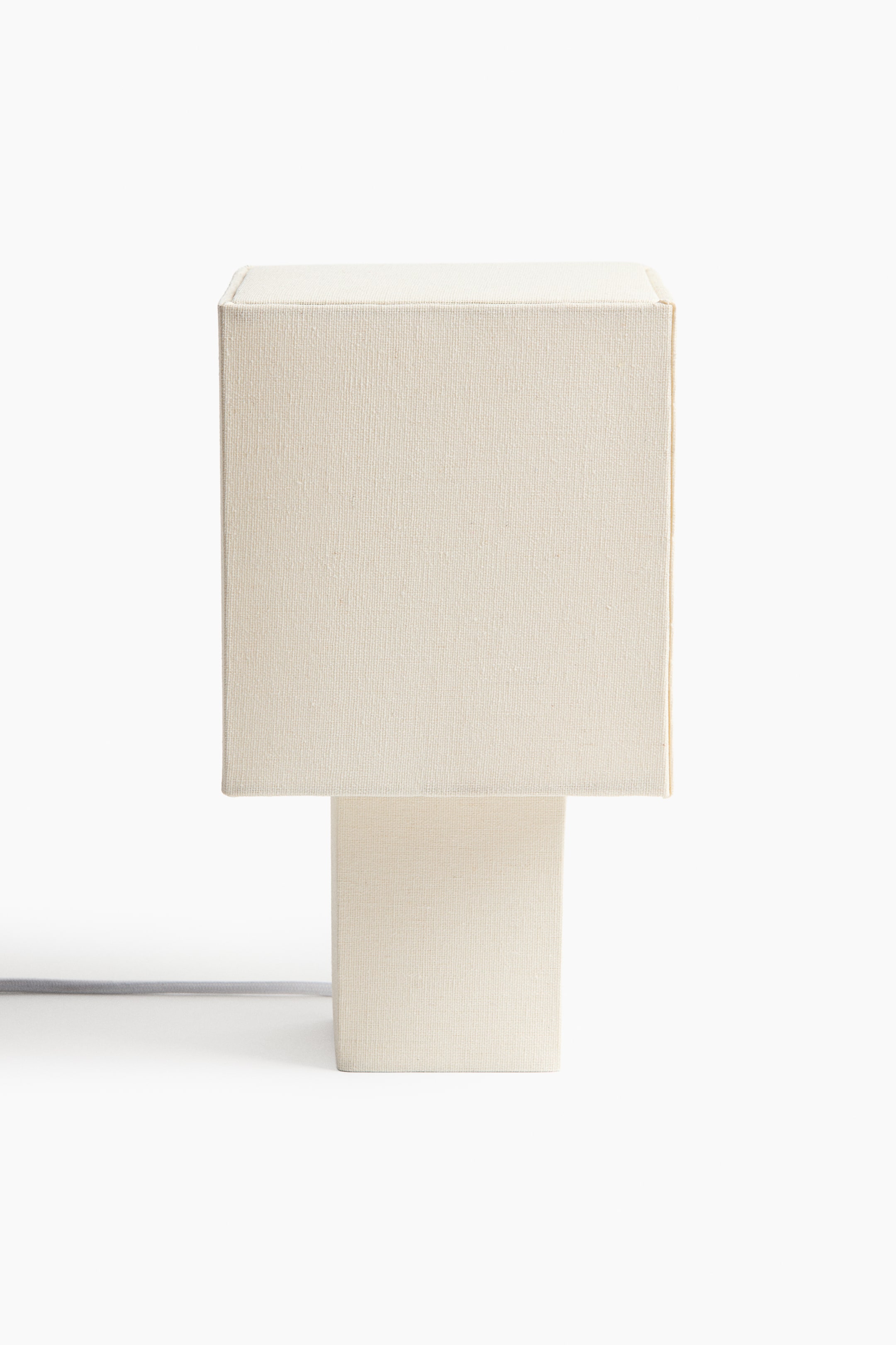 Agrandir l'image: Une lampe de couleur crème est orientée directement vers l’avant, présentant un abat-jour rectangulaire recouvert de tissu, placé au-dessus d’un socle rectangulaire plus étroit également recouvert de tissu.