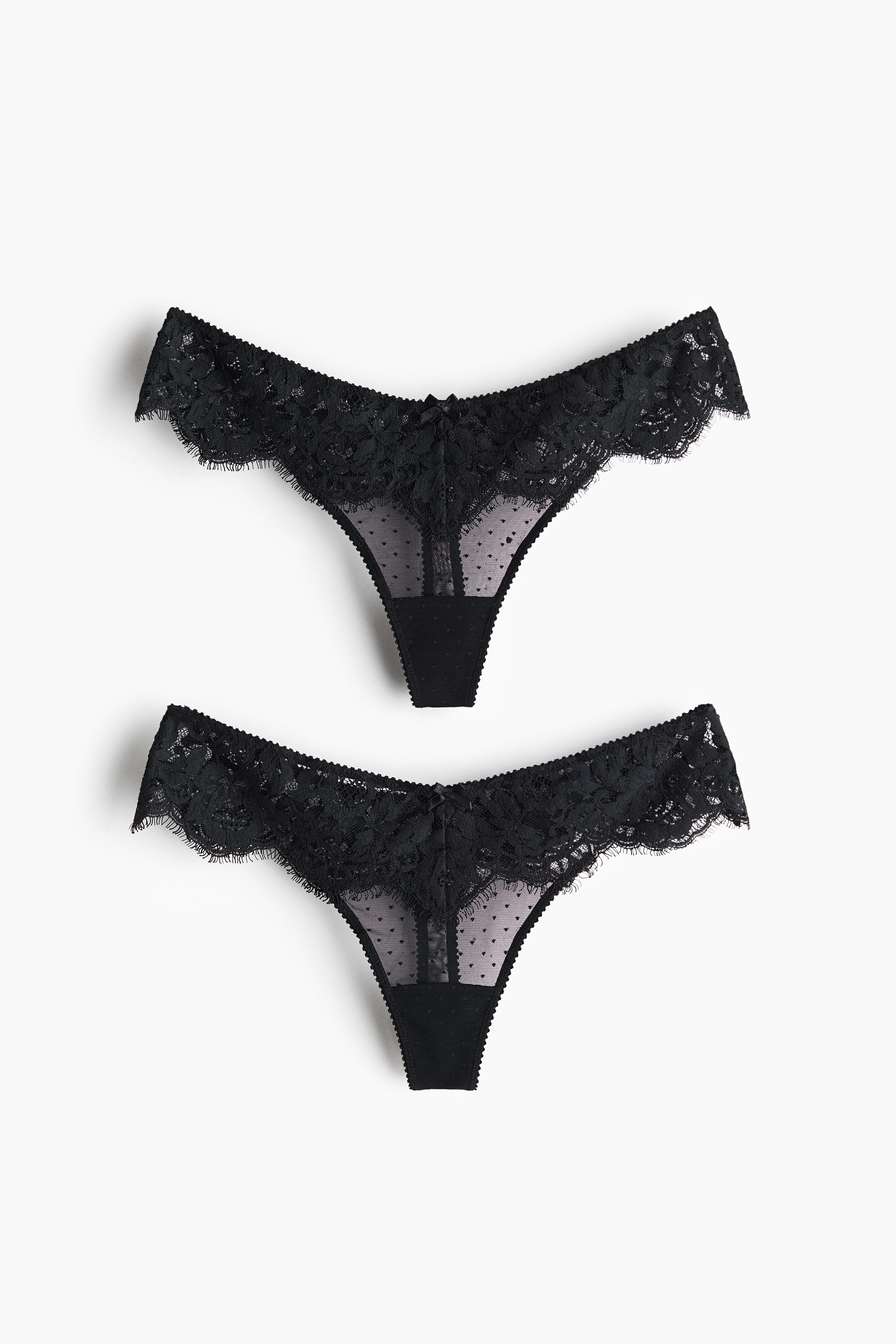 A black lace and mesh G-string with a low-rise waist is being worn by the model, styled with a matching black lace bra. The model is posing with arms raised behind the head, showcasing the side profile and fit of the underwear.