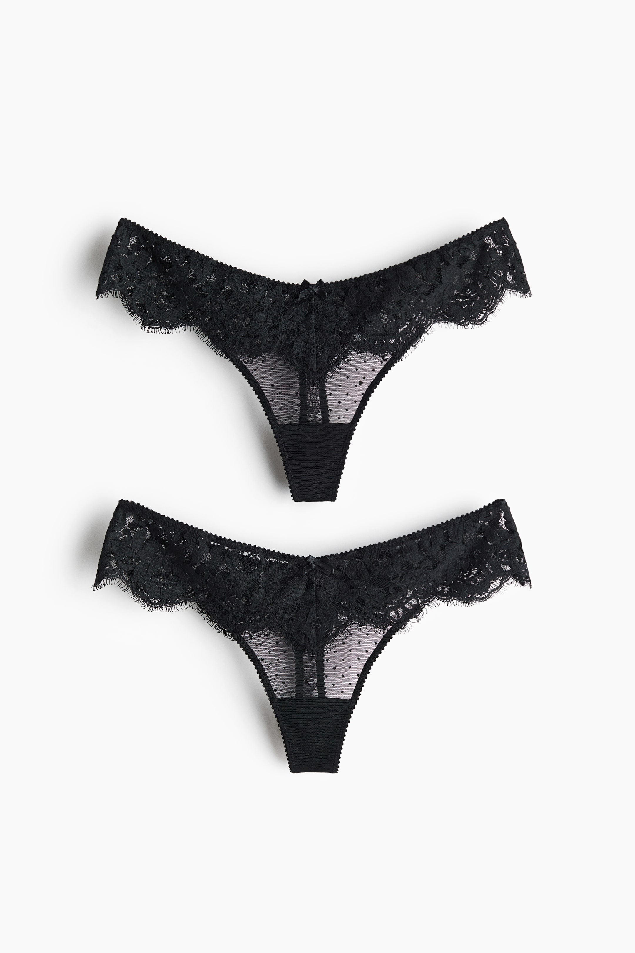 View larger image: Two black thongs are shown from the front, each featuring a scalloped lace waistband. The thongs are designed with a sheer mesh front panel adorned with small black polka dots and a small black bow at the centre.