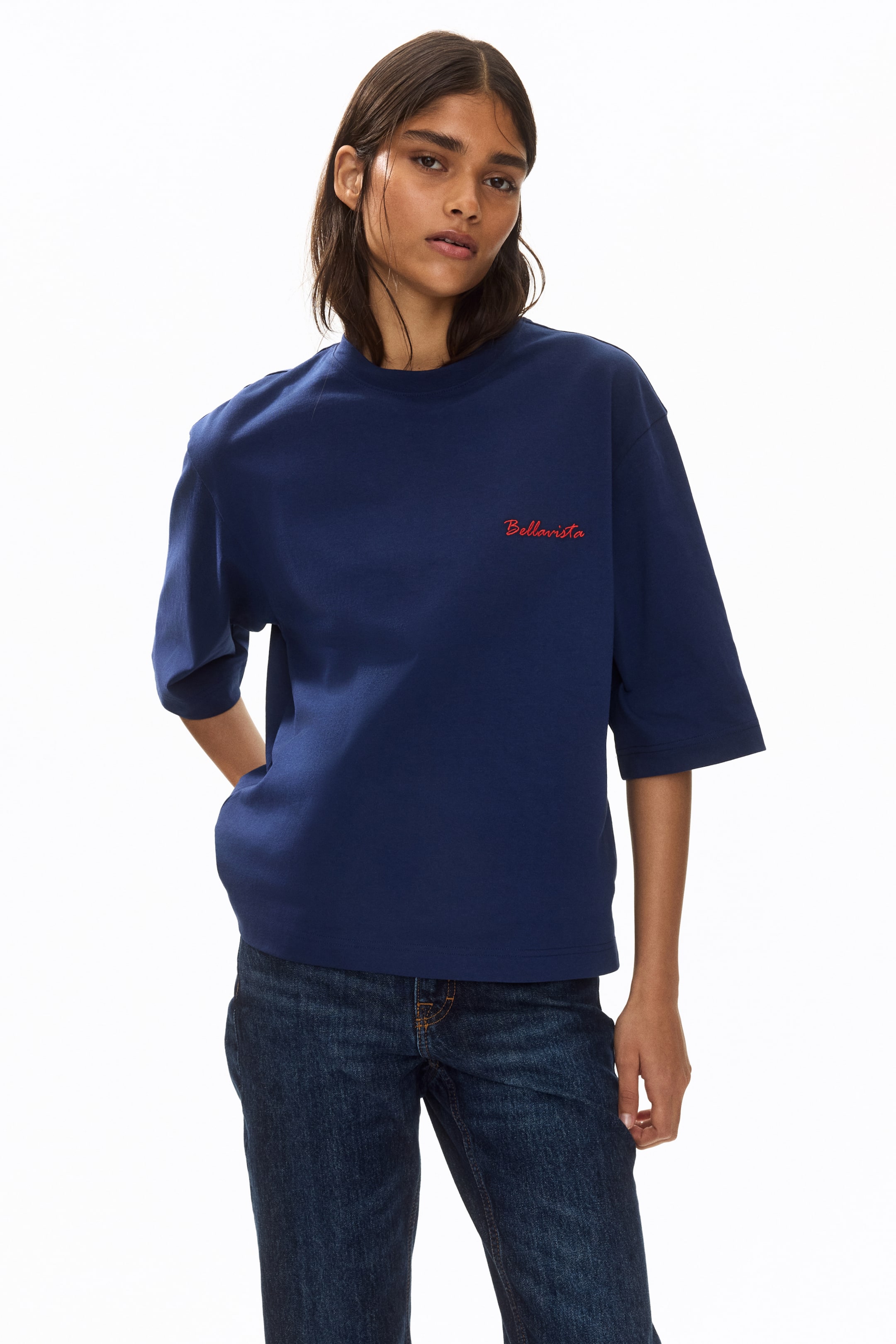 View larger image: The model is seen wearing an oversized navy blue t-shirt crafted from soft cotton jersey. This piece features a boxy silhouette with a round, rib-trimmed neckline, dropped shoulders, and elbow-length sleeves. The straight-cut hem complements the red "Bellavista" text embroidered on the chest. Styled effortlessly, the model places one hand in a jean pocket, pairing the t-shirt with dark wash jeans.