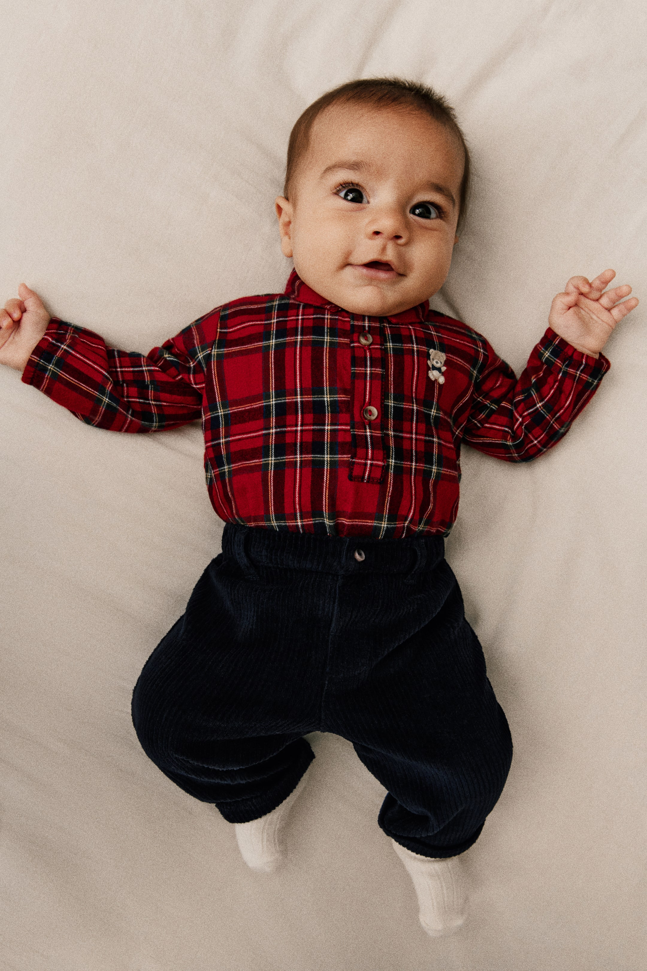 Agrandir l'image: Un ensemble de vêtements pour bébé est présenté de face, composé d’une combinaison en flanelle à carreaux rouge, noire et blanche, associée à un pantalon en velours côtelé bleu marine foncé. La combinaison présente un col rabattu, une patte de boutonnage avec un petit détail en forme d’ourson, ainsi que des manches longues avec poignets élastiqués, tandis que le pantalon affiche une finition texturée côtelée avec une fermeture à bouton.