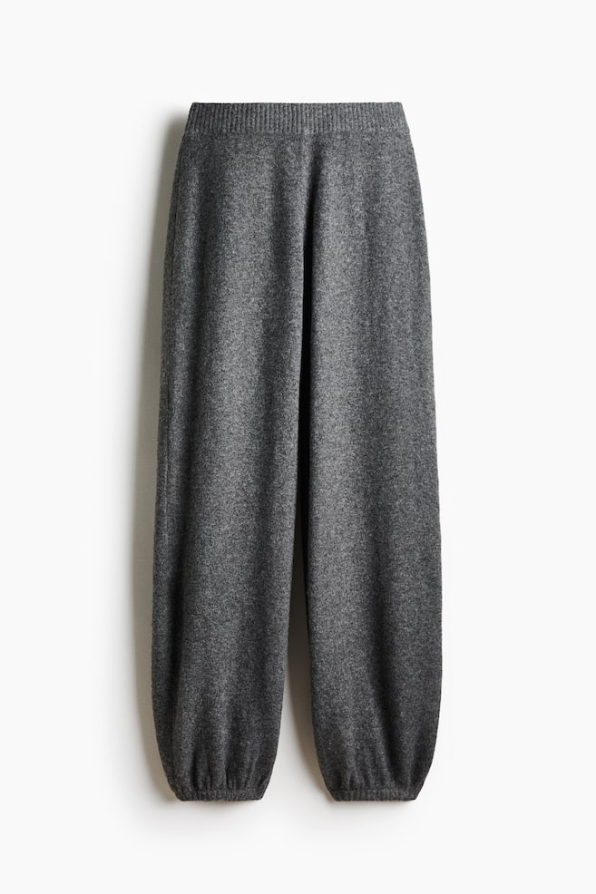 These grey knit trousers are displayed in a front-facing view, featuring a thick ribbed waistband and a relaxed, wide-leg silhouette. The legs taper down into gathered, ribbed ankle cuffs, showcasing the textured, heathered grey fabric. - 1