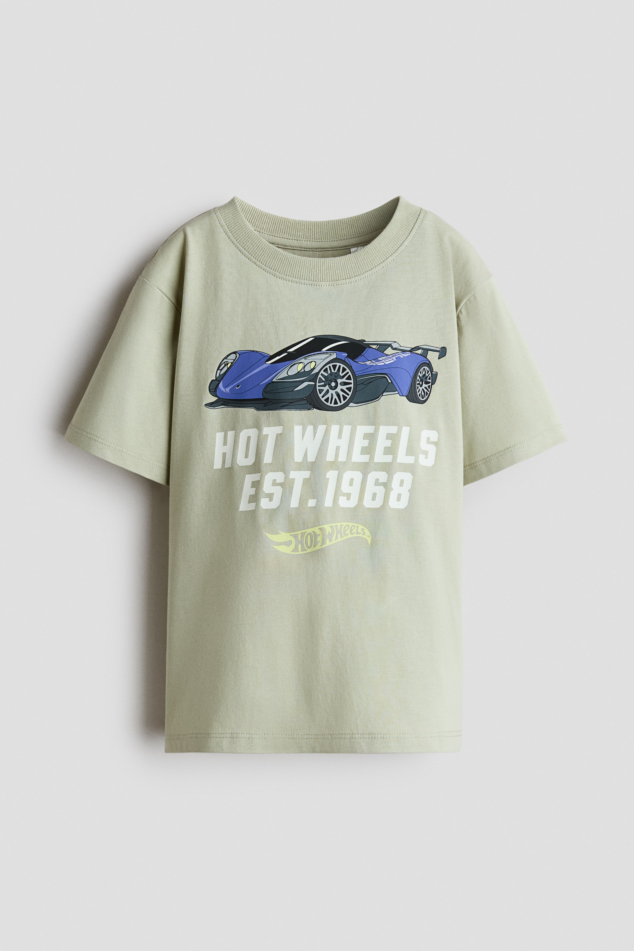 View larger image: A soft, light green printed cotton jersey T-shirt is shown from the front, featuring a rib-trimmed crew neckline and gently dropped shoulders. The front displays a blue and black race car graphic above white text reading 'HOT WHEELS EST. 1968' and a yellow Hot Wheels logo.