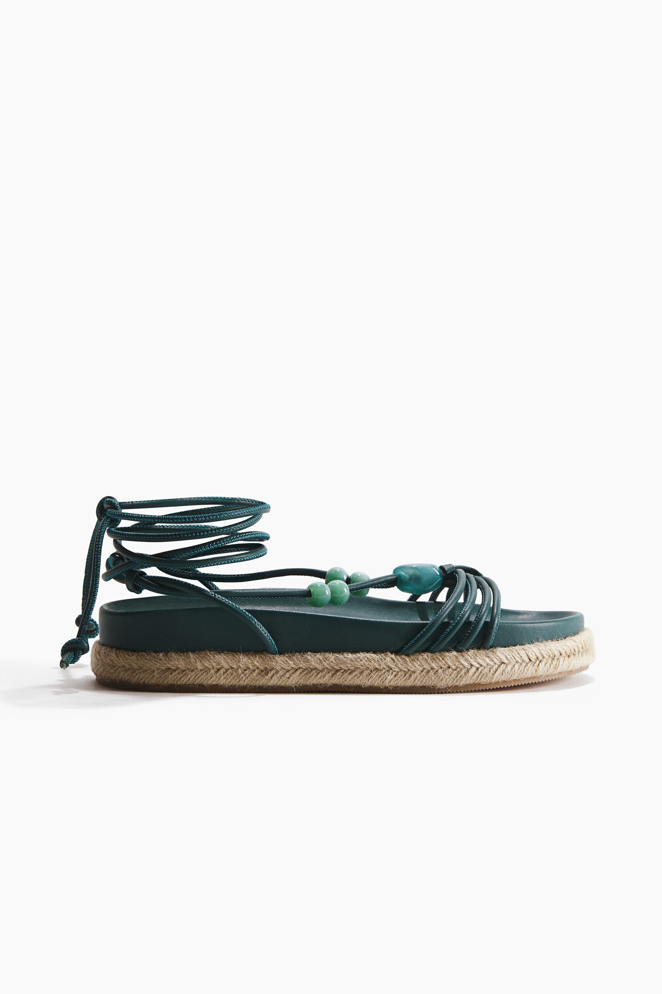 Visualizza l'immagine ingrandita: Il sandalo verde scuro presenta un plantare piatto e una spessa suola a piattaforma, con una treccia in espadrille dai toni naturali che avvolge i lati. Molteplici cinturini stretti verde scuro attraversano l'area delle dita, adornati con perline verde chiaro e verde teal scuro, mentre lunghi lacci si estendono dai lati, formando un'allacciatura alla caviglia.