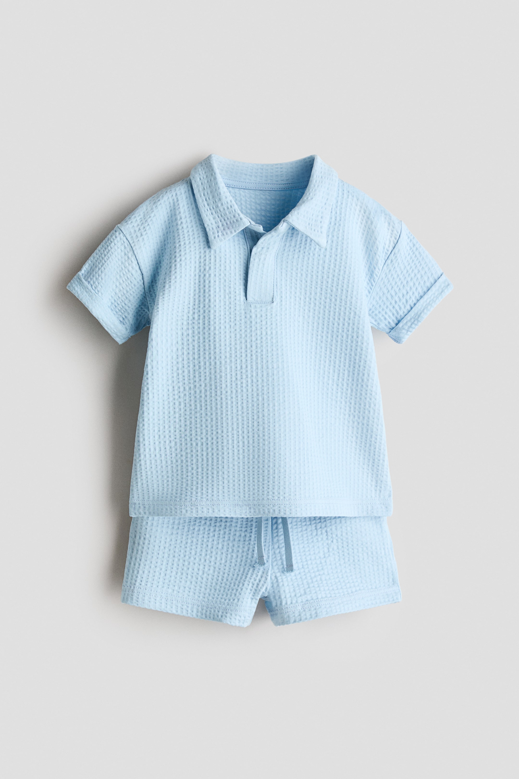 View larger image: A light blue waffle-knit two-piece set is shown from the front, featuring a short-sleeved polo top with a collar, a v-neck placket, and rolled cuffs, paired with matching shorts that have a drawstring waist.