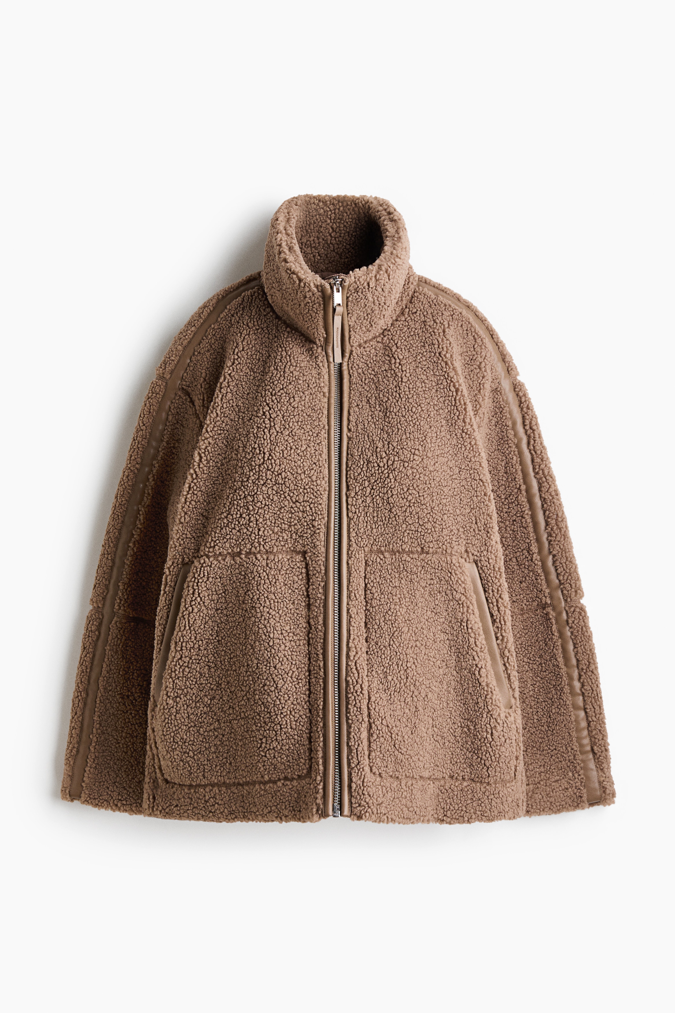 En oversized teddyjacka i ljusbrun fÃ¤rg bÃ¤rs av modellen och har en hÃ¶g funnelkrage, stora sidofickor och en central dragkedjestÃ¤ngning med integrerad babybÃ¤rpanel. Modellen bÃ¤r ett baby som Ã¤r sÃ¤krat i den frÃ¤mre panelen samtidigt som hen stÃ¶der barnet med en hand.