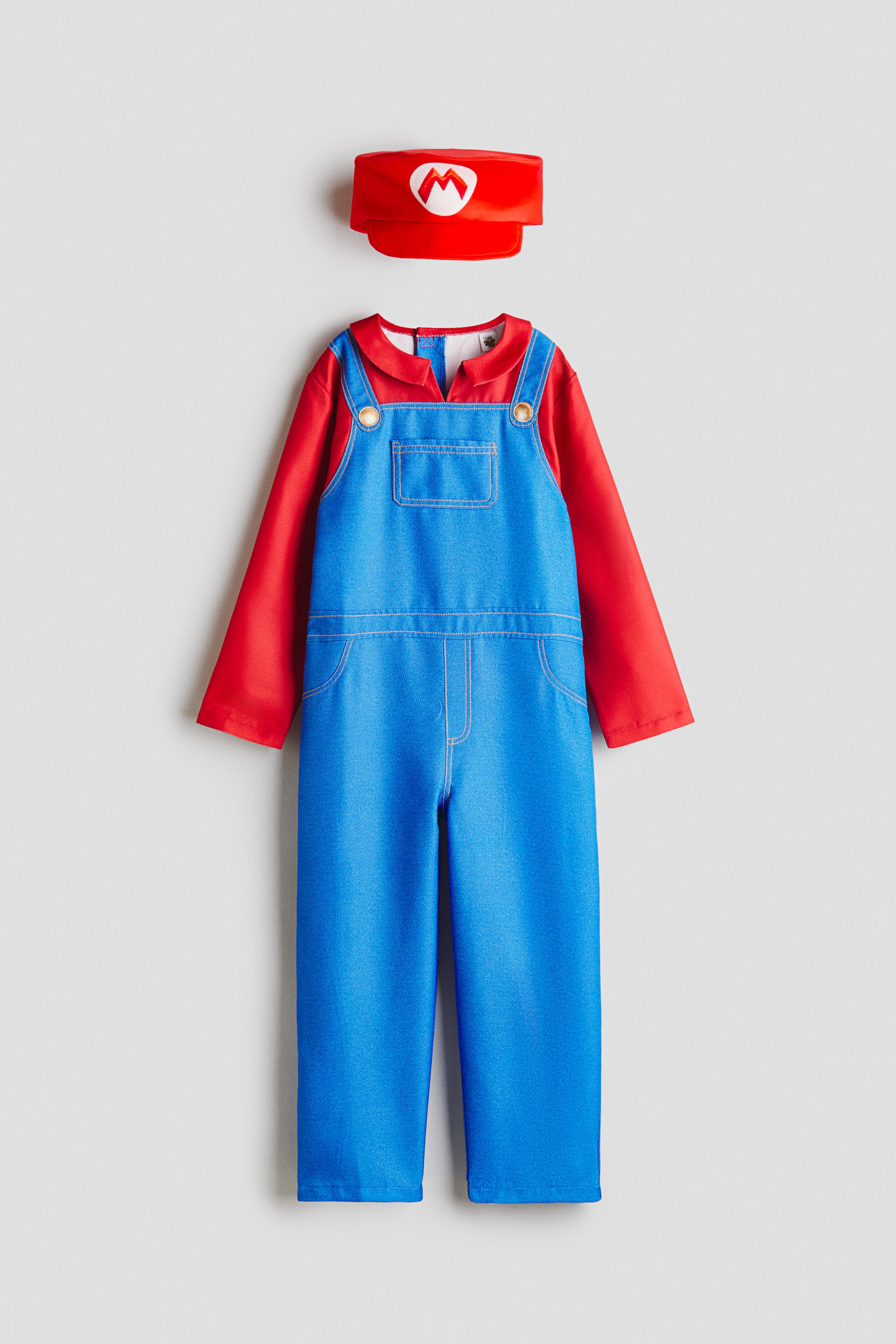View larger image: A front-facing costume features a red long-sleeved shirt layered under blue overalls, detailed with gold-toned buttons on the straps, a stitched front bib pocket, and white seam lines. A matching red hat, with a white 'M' emblem on the front, is positioned above the costume.