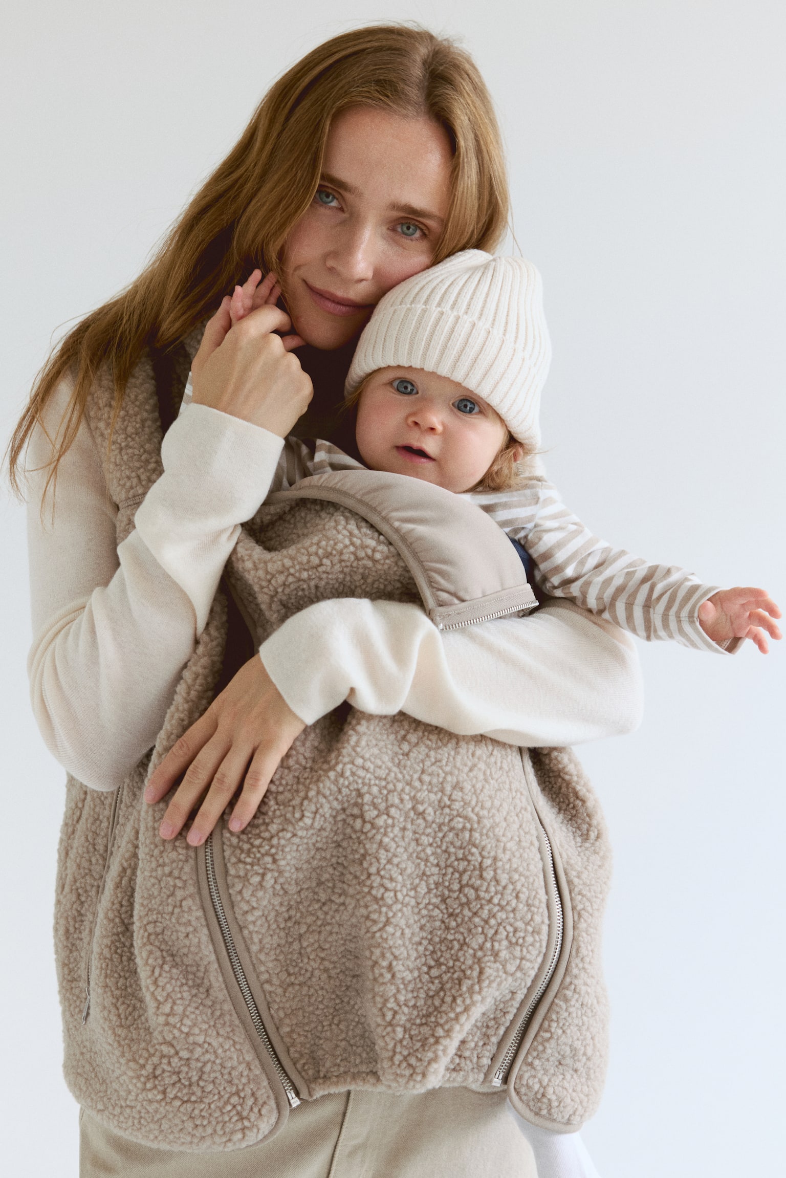 A beige sherpa vest is featuring a full-length metal zipper down the front and a stand-up collar. Two zippered pockets are visible on the lower front, and matching fabric trim is bordering the zipper, collar, armholes, and hem. - 1