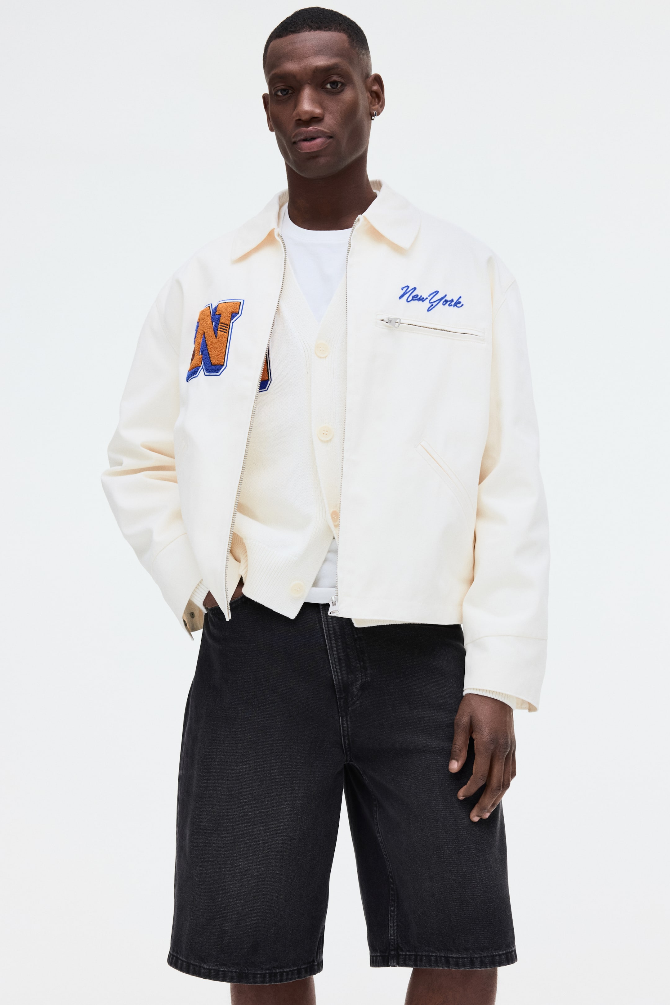Grösseres Bild ansehen: Eine cremefarbene Jacke mit spitzem Kragen, durchgehendem Frontreißverschluss und gerippten Bündchen und Saum. Sie zeigt einen großen orange-blauen "N"-Aufnäher auf der linken Brust, eine blaue "New York"-Stickerei auf der rechten Brust, schräge Leistentaschen und eine Brusttasche mit Reißverschluss und wird offen über einem cremefarbenen Cardigan getragen.