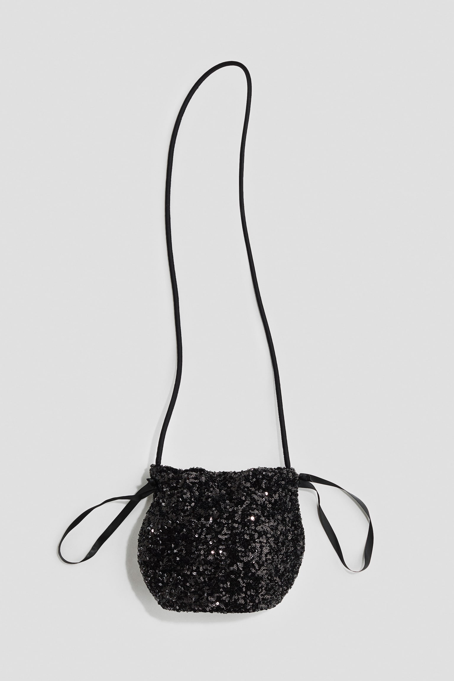 A small, black sequined pouch bag is shown from the front, featuring a thin black cord shoulder strap. The bag highlights a gathered drawstring closure at the top with black satin ribbons looping out on both sides, and its entire exterior is covered in shimmering black sequins.