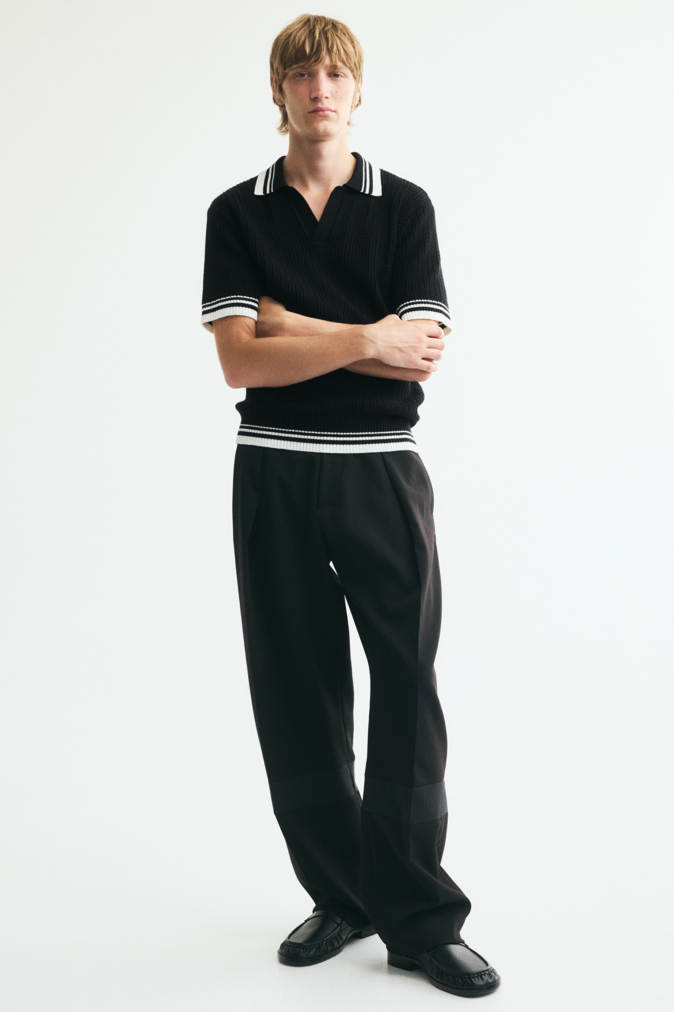 View larger image: A black, textured-knit short-sleeve polo shirt with contrasting white striped trims on the open collar, cuffs, and hem is worn by the model. The model is standing with arms crossed, pairing the shirt with wide-leg black trousers and black shoes.