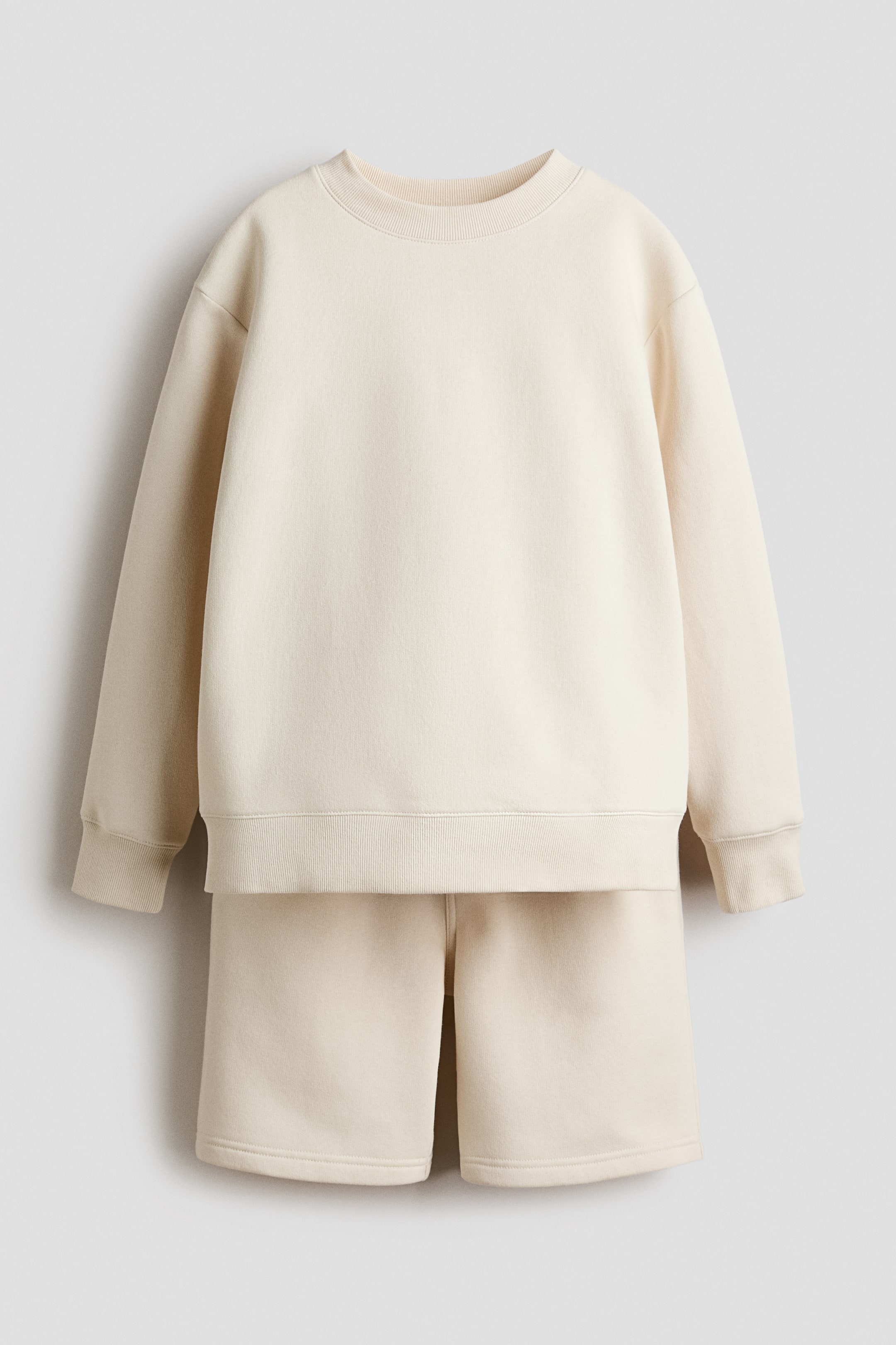 Zobrazit větší obrázek: Zepředu je představen světle béžový set mikiny, který obsahuje dlouhý rukáv s kulatým výstřihem, spadlými rameny a žebrovanými detaily na výstřihu, manžetách a lemu. Odpovídající kraťasy po kolena mají elastický pas.