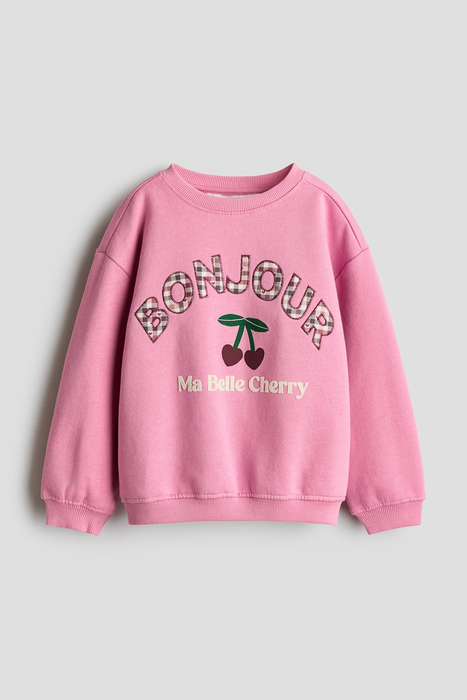 A pink crew-neck sweatshirt is shown from the front, featuring dropped shoulders, ribbed cuffs, and a ribbed hem. The front displays an arched 'BONJOUR' applique in a brown-and-white gingham pattern, positioned above a cherry graphic and the embroidered text 'Ma Belle Cherry'.