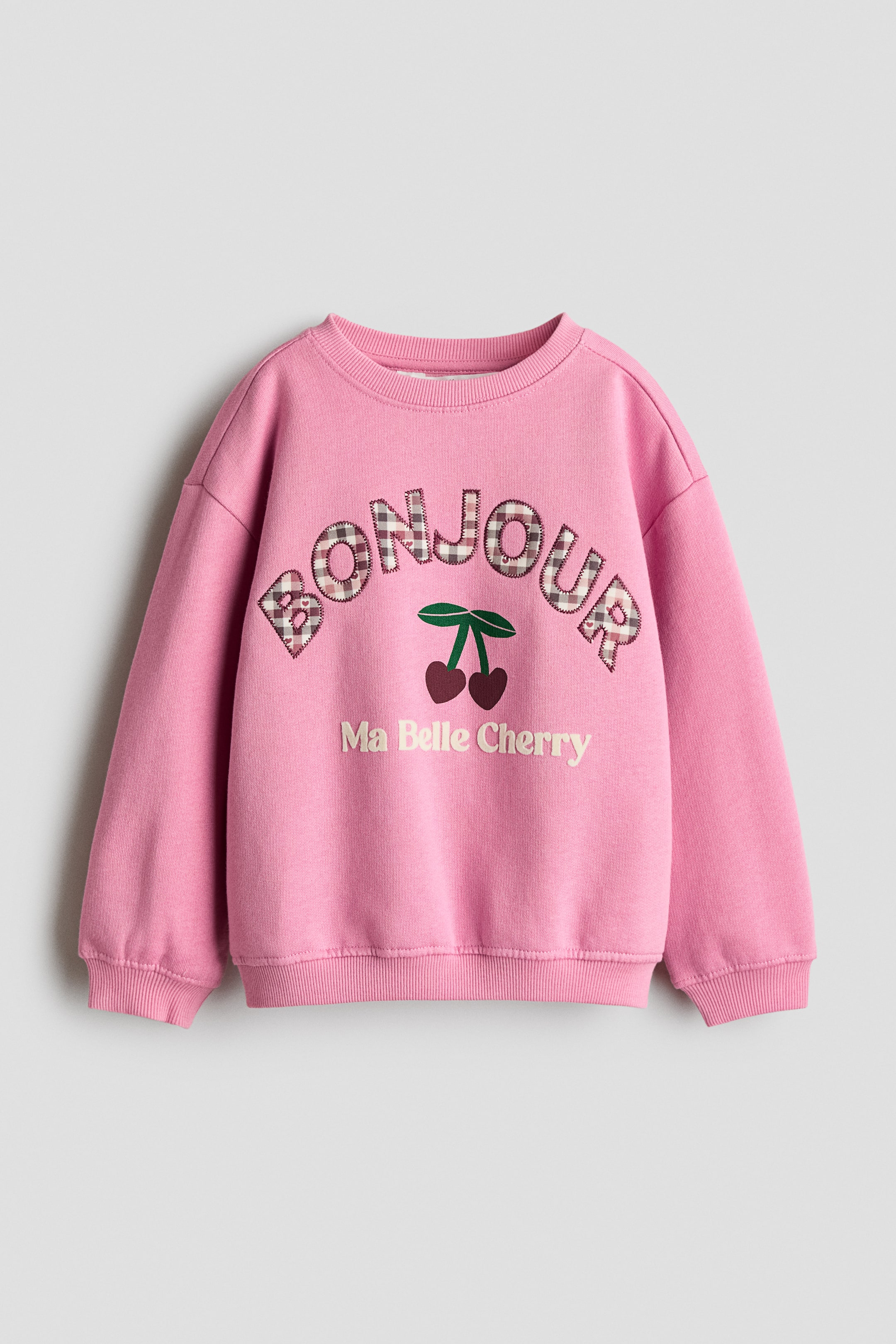 View larger image: A pink crew-neck sweatshirt is shown from the front, featuring dropped shoulders, ribbed cuffs, and a ribbed hem. The front displays an arched 'BONJOUR' applique in a brown-and-white gingham pattern, positioned above a cherry graphic and the embroidered text 'Ma Belle Cherry'.