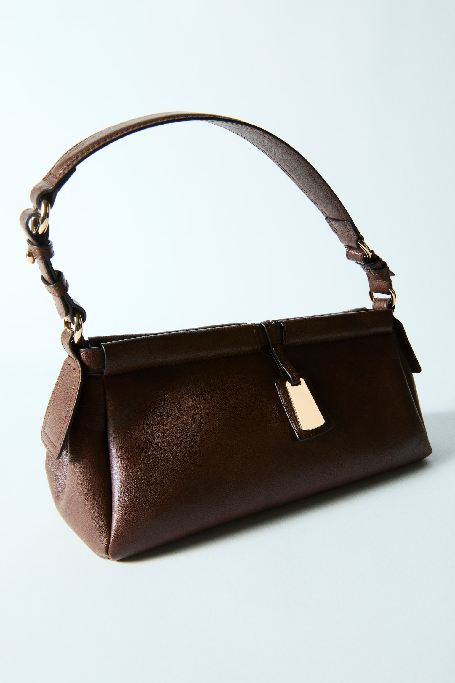 A dark brown shoulder bag is showcasing its front view, boasting a smooth texture and a slim, adjustable strap adorned with gold-toned fittings. A rectangular gold-toned charm dangles from the central flap closure. - 1